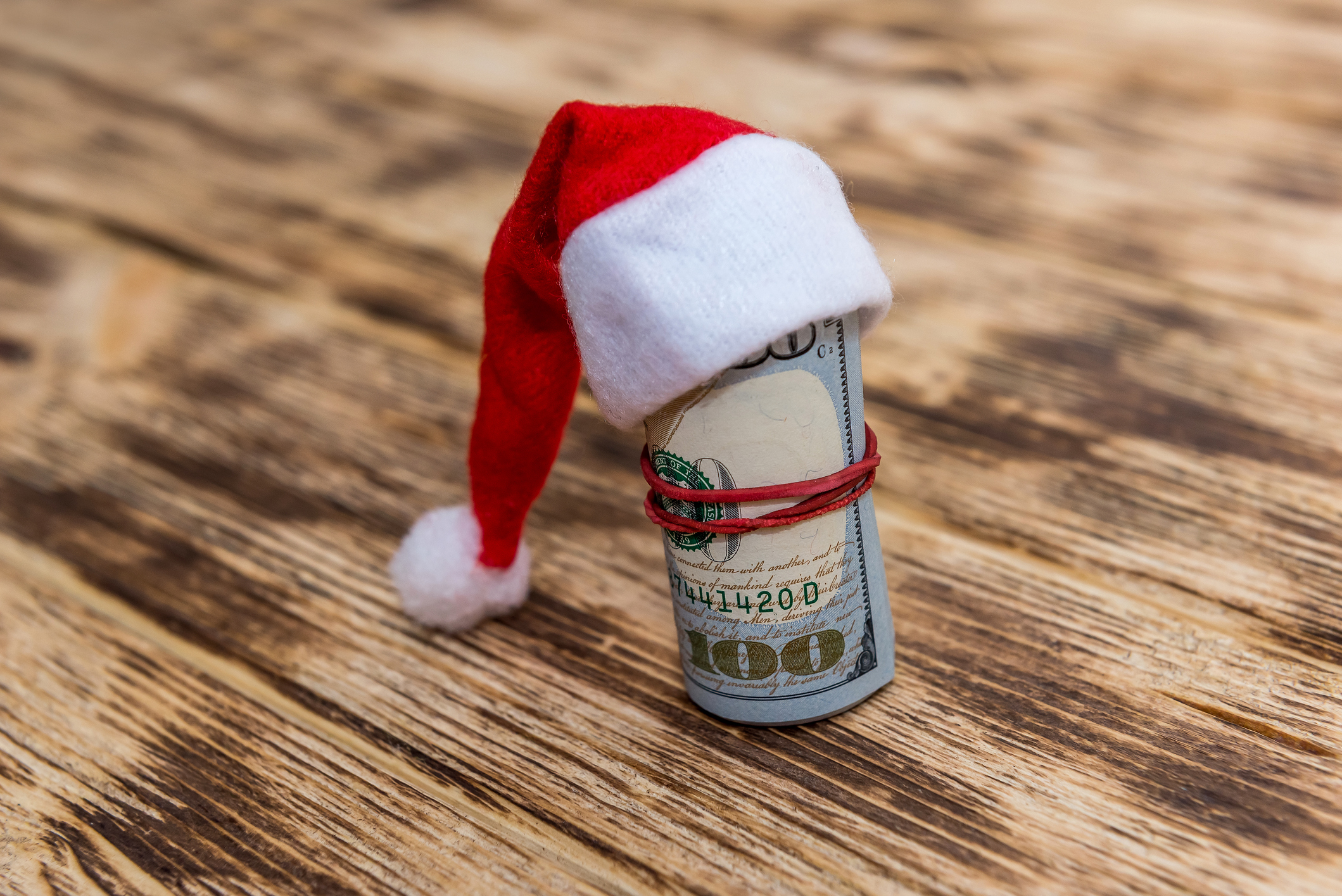 a $100 bill rolled up and wearing a Santa hat on a wood surface