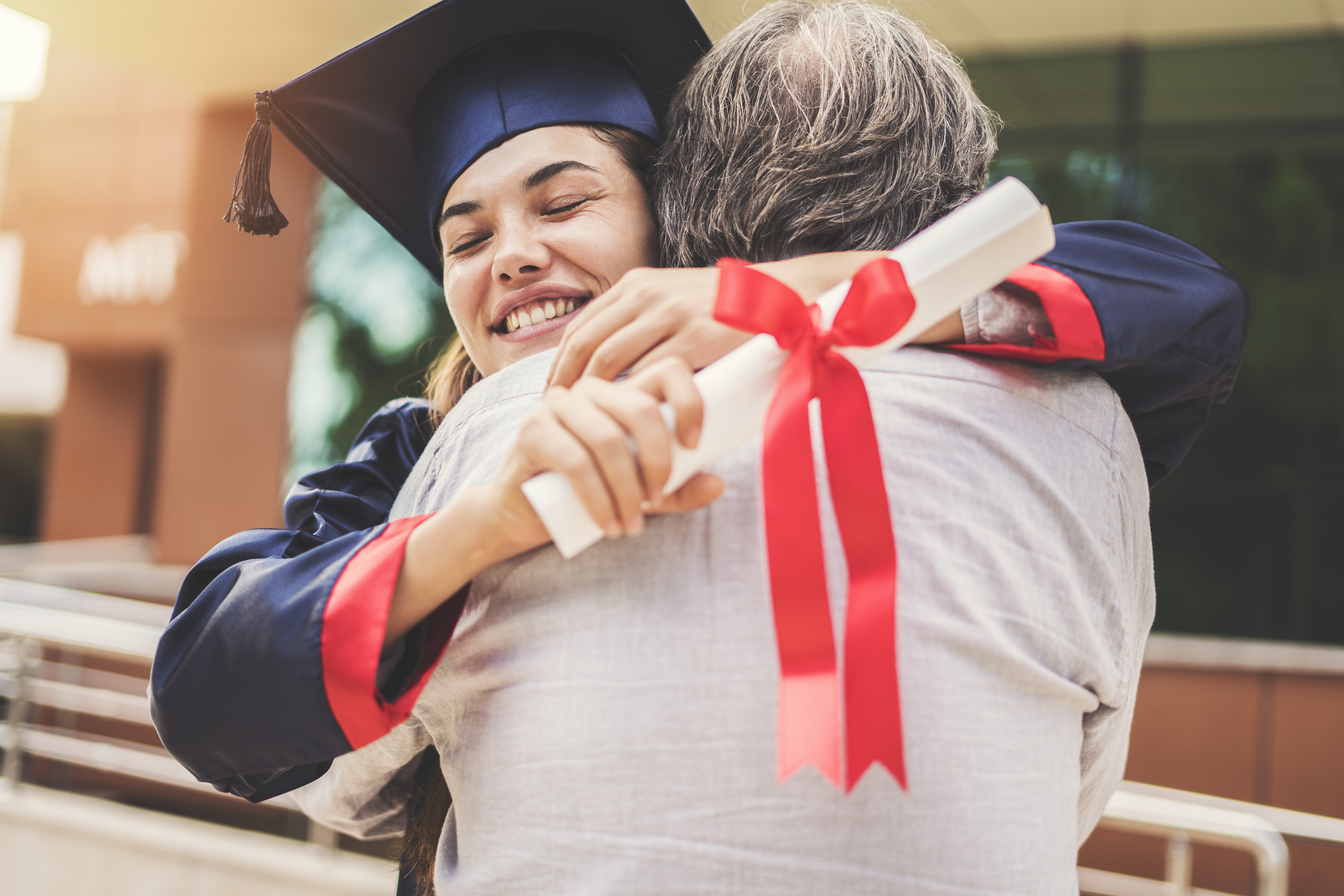 A young woman in a cap and gown holding a diploma hugs an older person.