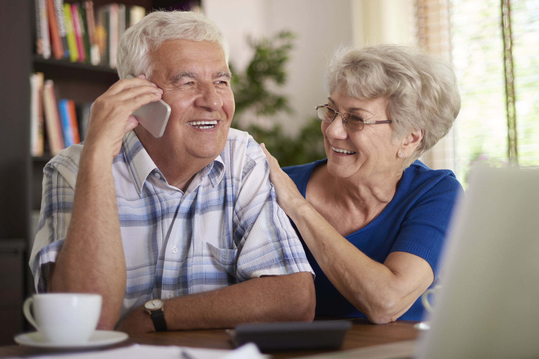 Older woman putting hand on older man's arm while he talks on the phone; both are smiling