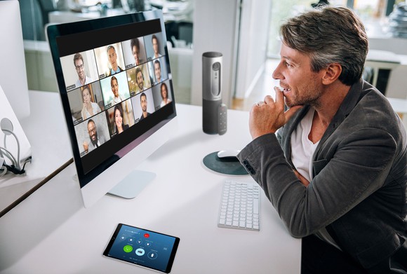 A man at his desk looking at a computer screen showing the faces of 12 people. 