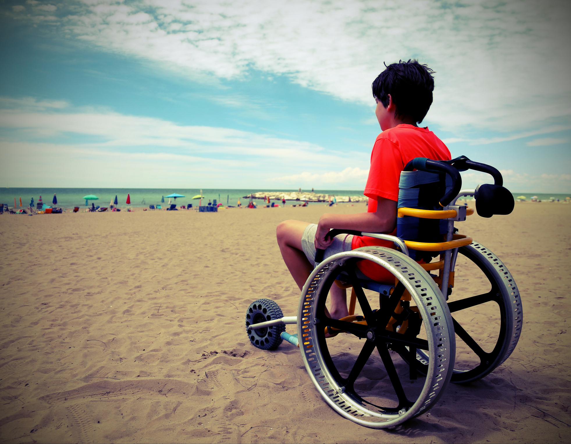 A young boy in a wheelchair that's on a sandy beach.