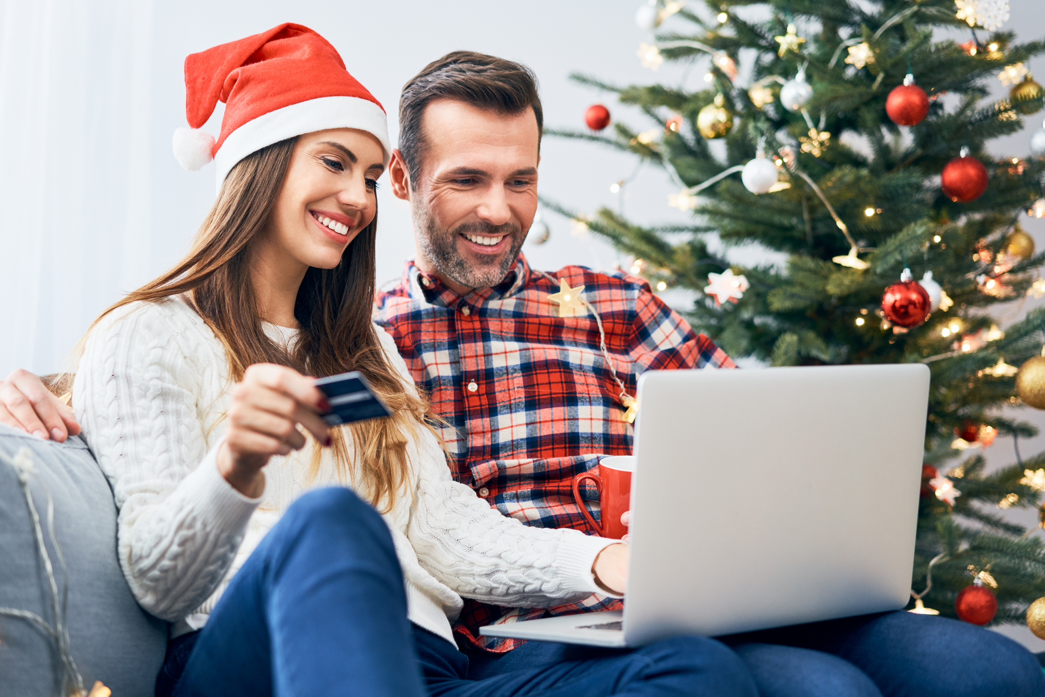 A woman in a Santa hat and a man sitting on a couch entering credit card data on a laptop with a Christmas tree in the background.