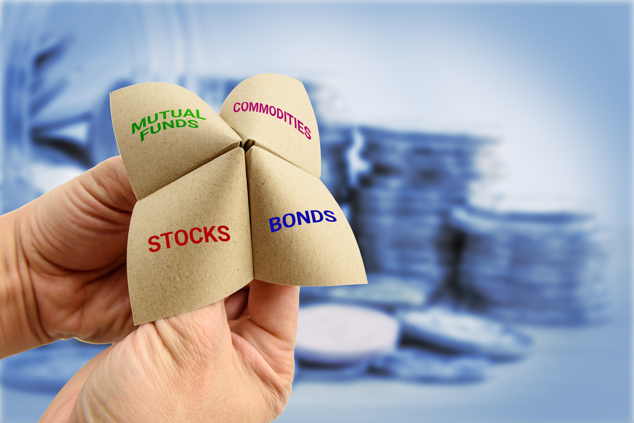 A child's folded paper finger toy is shown, with the words stocks, bonds, commodities, and mutual funds written on it.