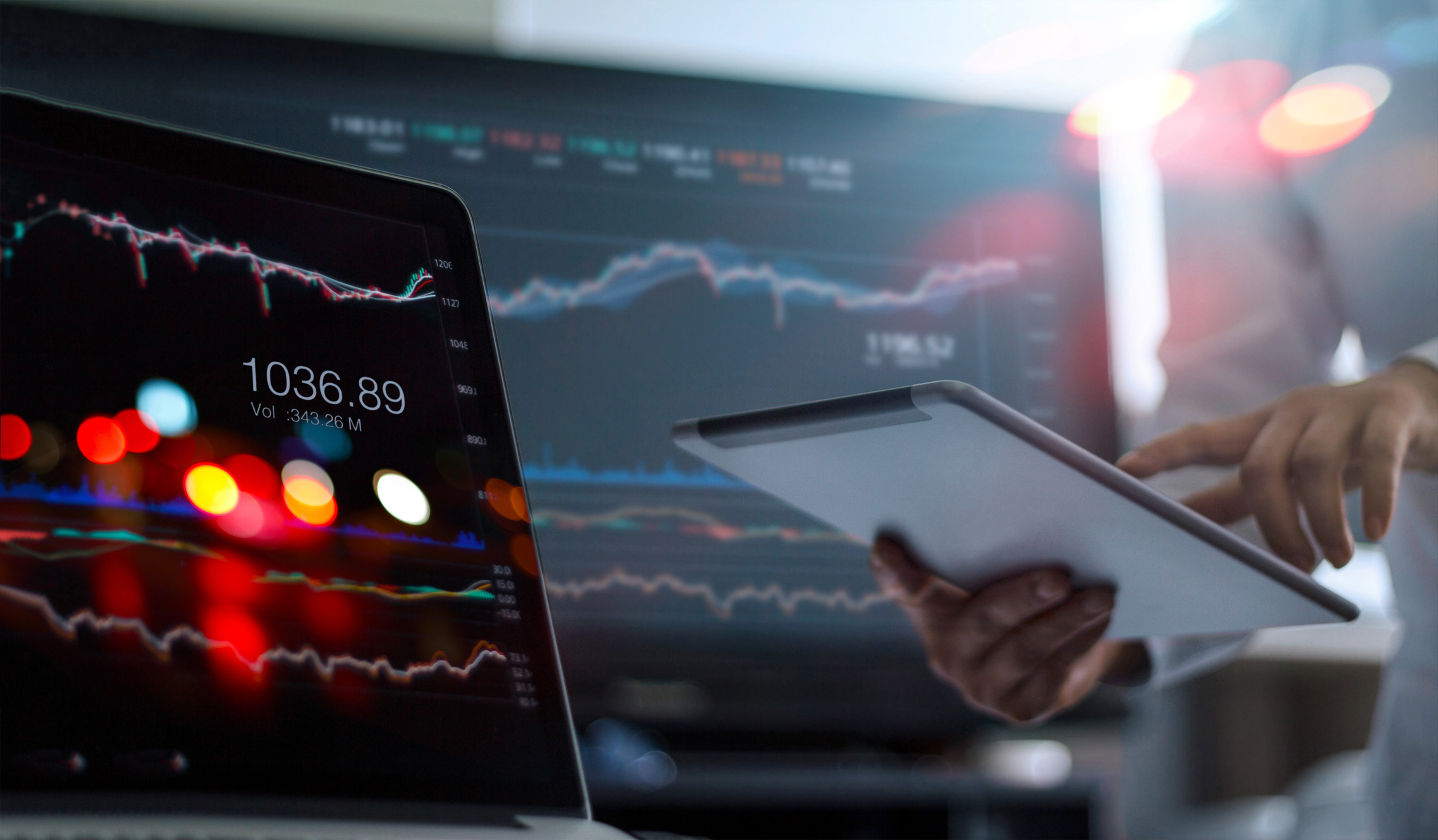 A man holding a tablet computer with computer screens displaying stock charts in the background.