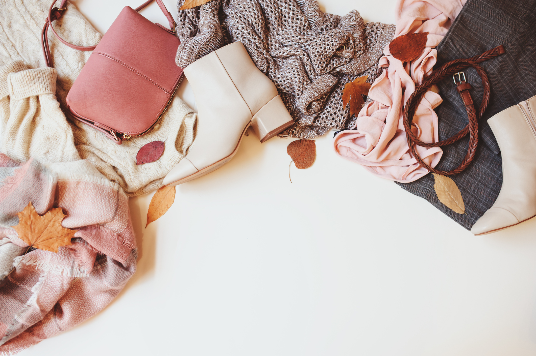 Pink and ecru women's ankle boots, handbag, scarf, and belt with fall leaves are spread on a cream-colored surface.