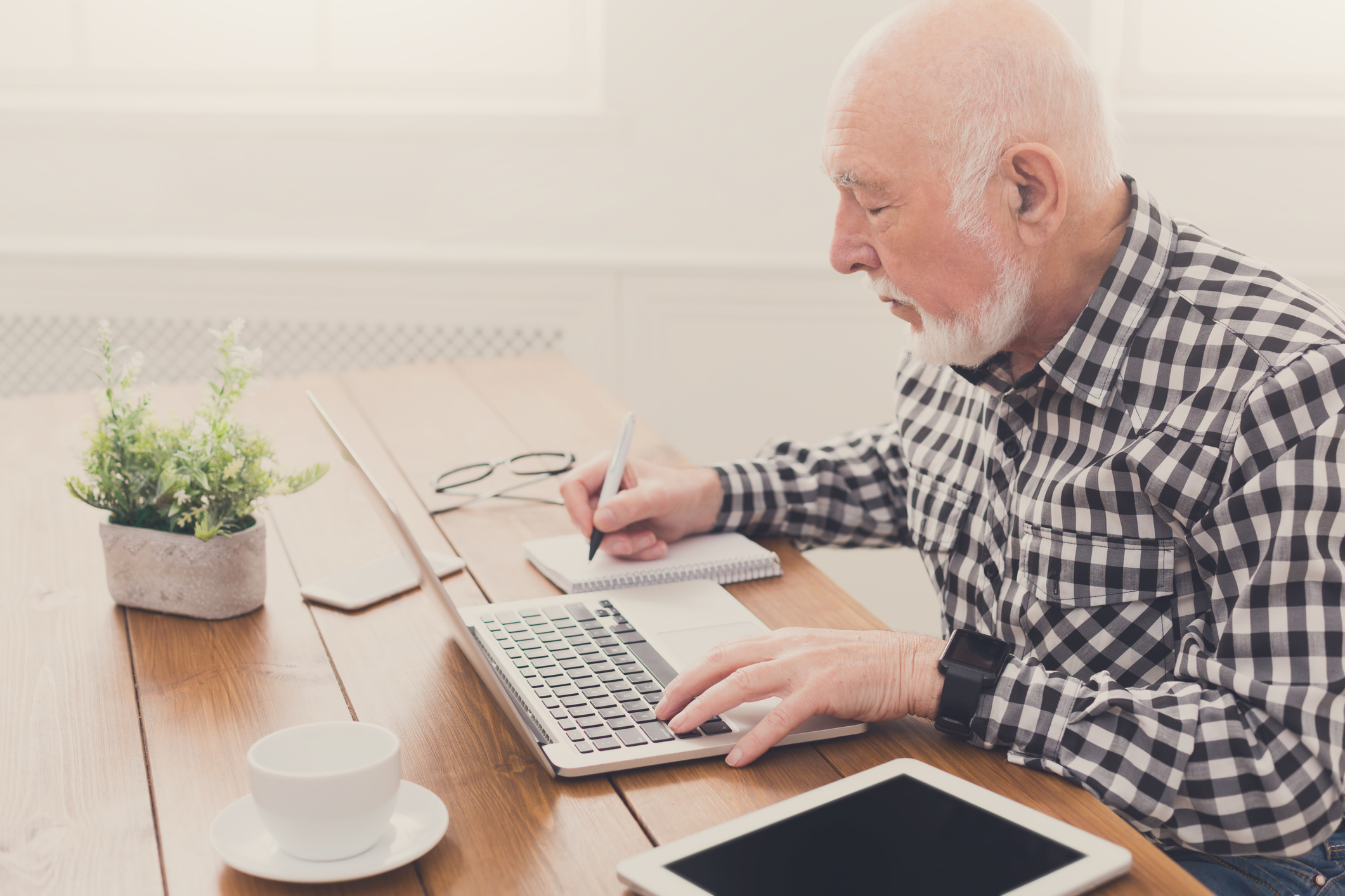Older man typing on laptop while writing on a notebook