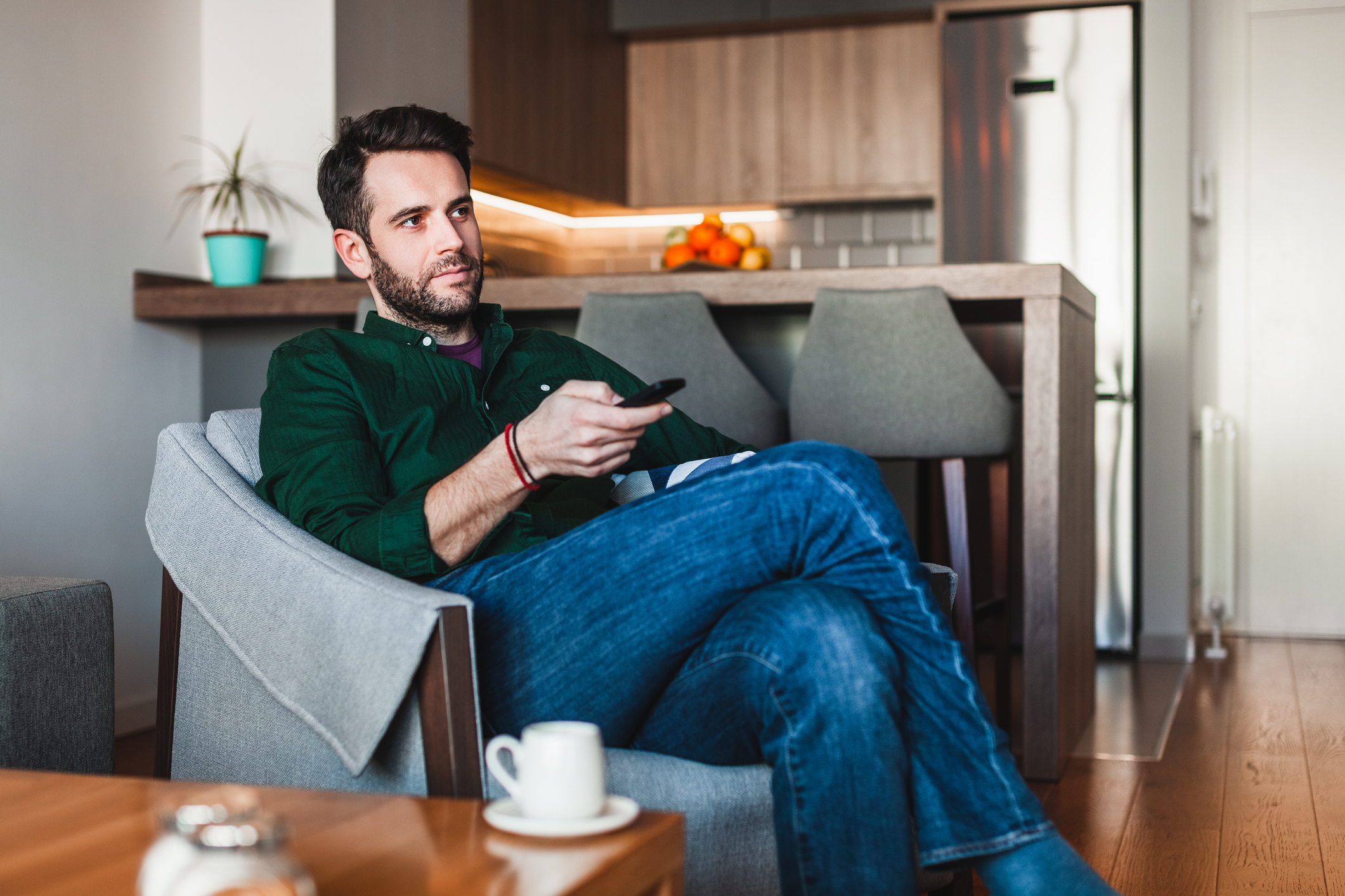 A man seated in a living room points a remote control at an unseen TV.