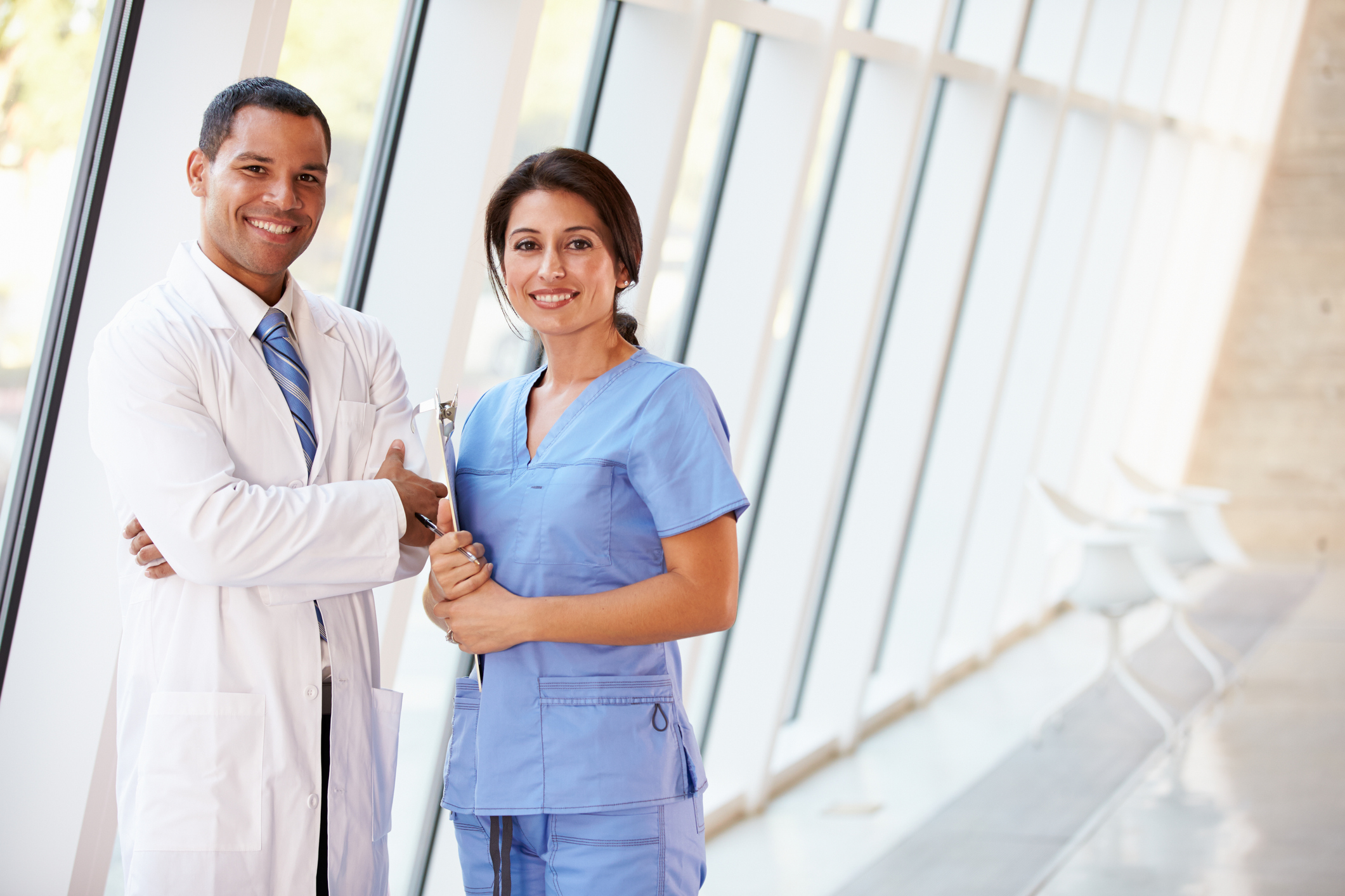 Male healthcare provider in white coat standing next to female healthcare provider in scrubs, both smiling.