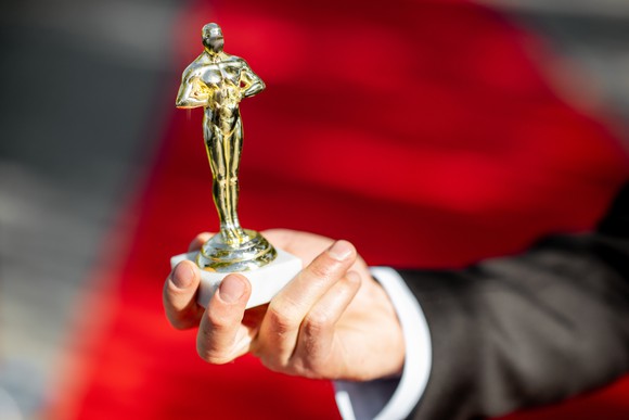 A hand holding up an unofficial copy of the famous Oscar statuette against the backdrop of a red carpet.