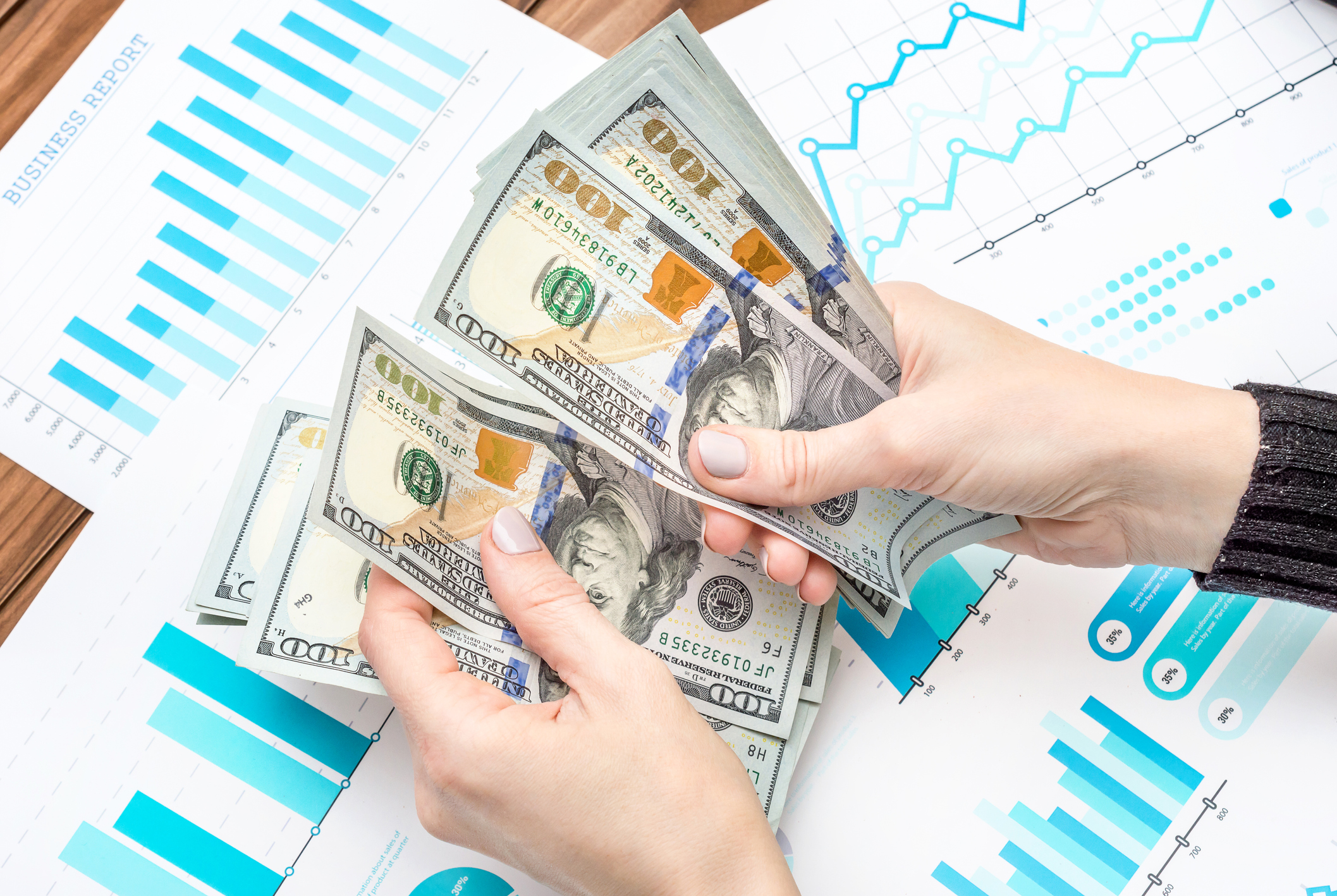 Two hands counting money over business charts on a desk.
