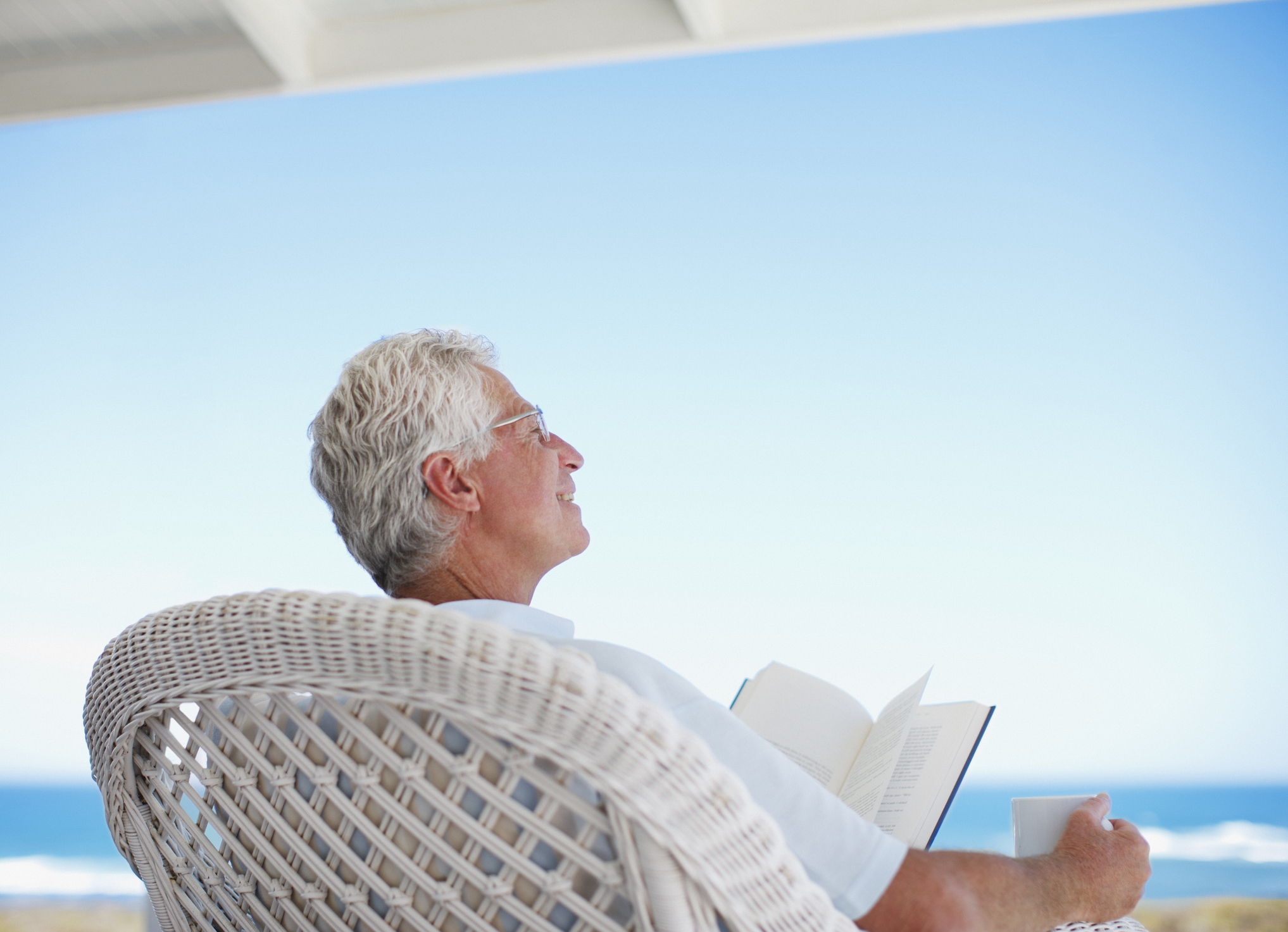 Older man in wicker chair reading book, holding mug, and facing the ocean