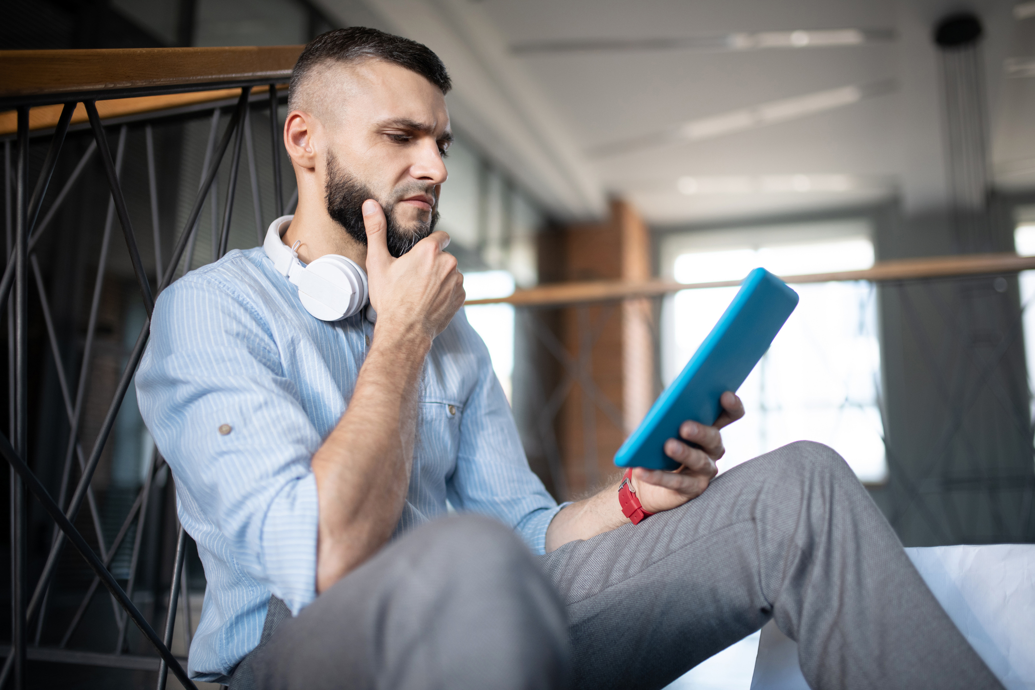 Young man sitting and stroking beard while holding tablet and sporting worried look on face