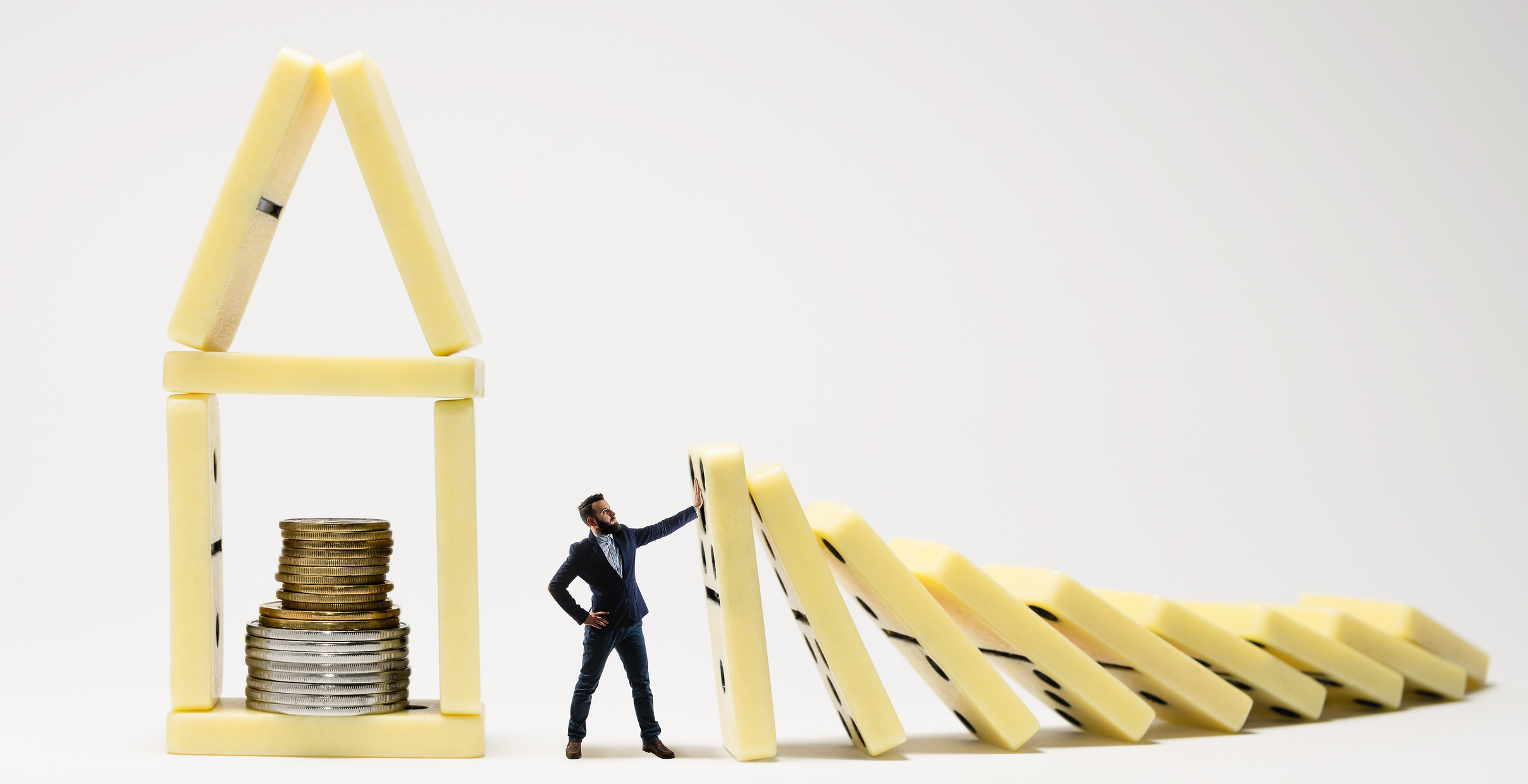 A man in a suit stops dominoes from knocking down a house made of dominoes with coins in it.