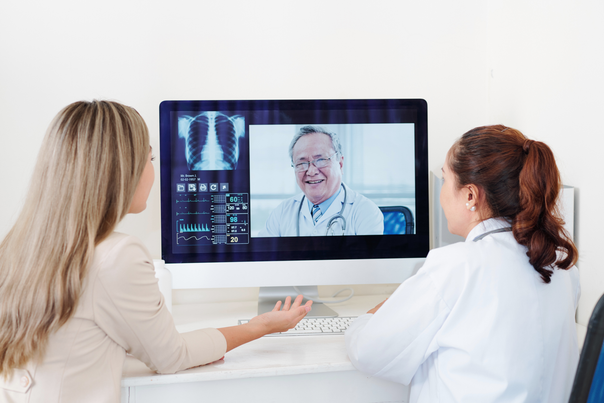 Two young women gather round a computer and speak with a doctor through a video conference via desktop.