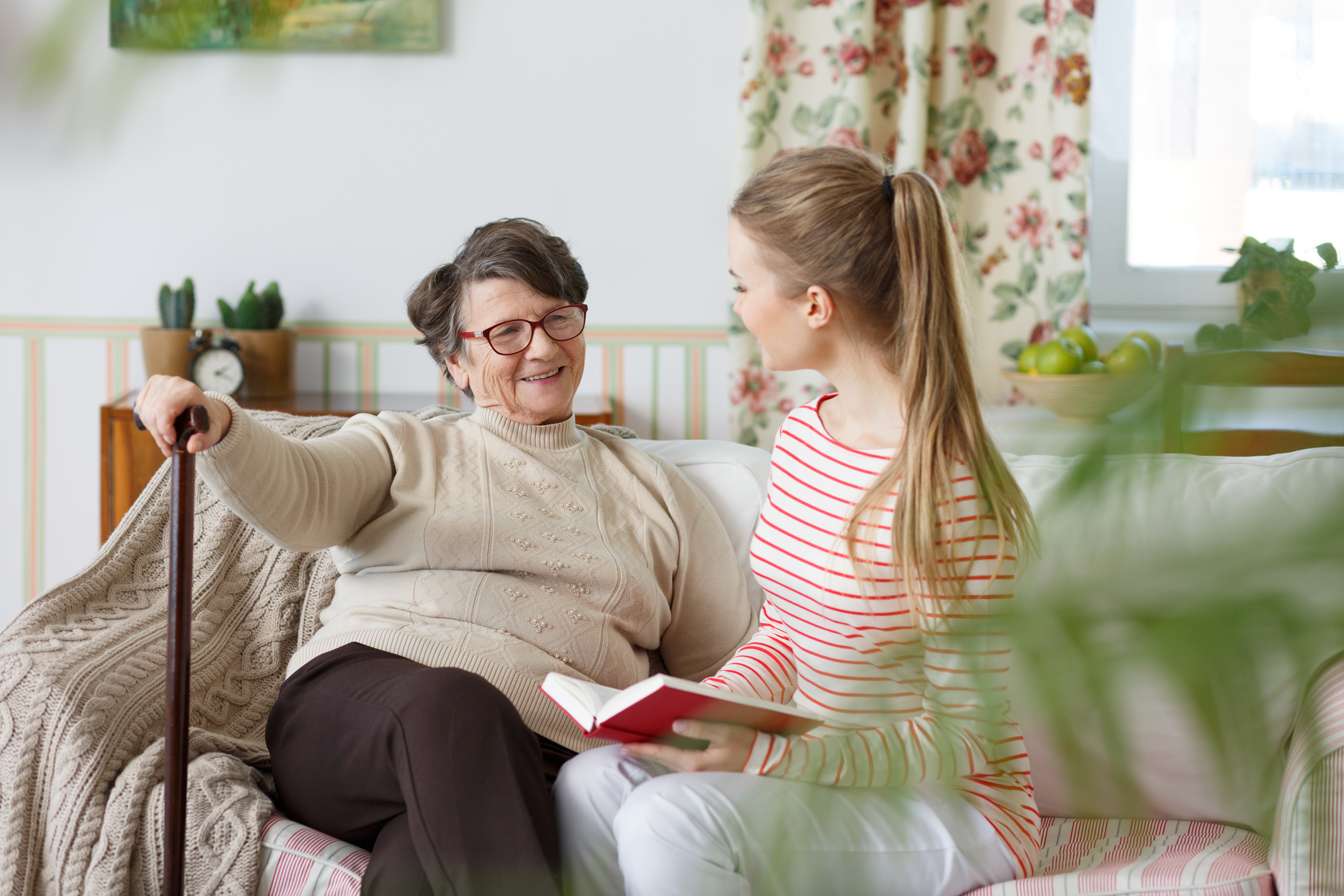 A young woman sits on a sofa next to a smiling older woman with a cane.