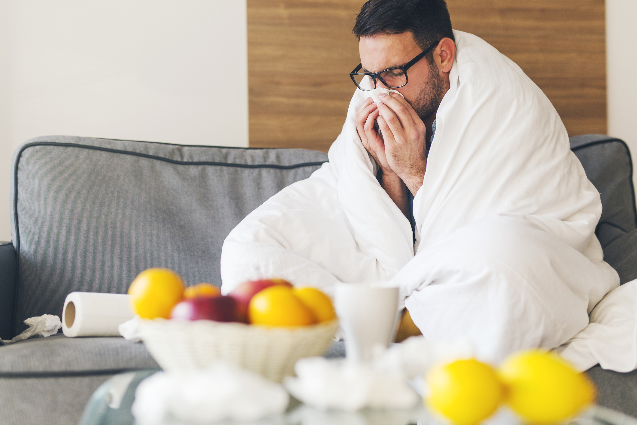 A man wrapped in a blanket sneezing into a tissue while sitting on a couch.