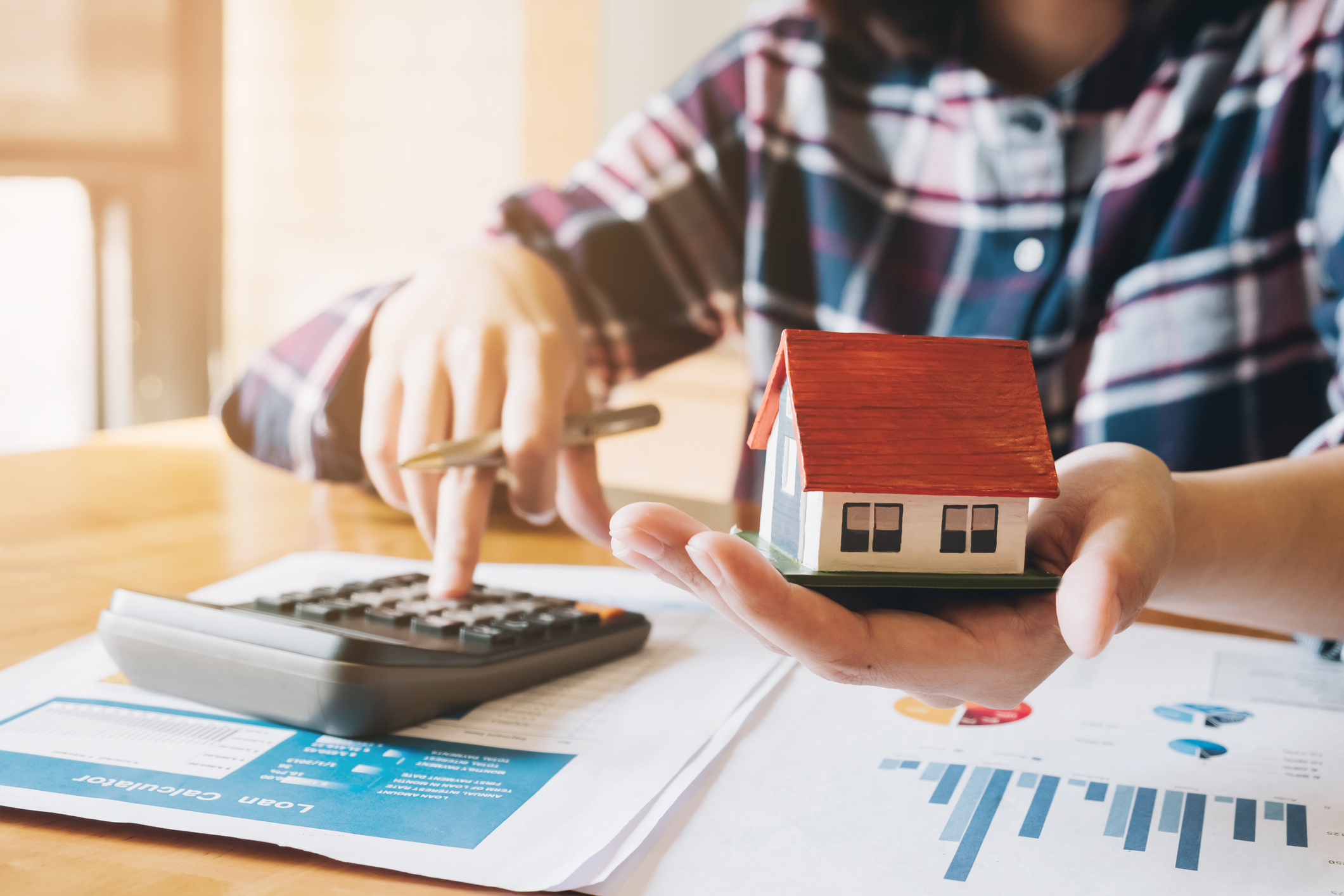 A woman holding a small model house works on a calculator with charts underneath it.