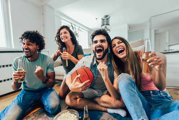 Twnety somethings root on a basketball game watching TV on the floor. 