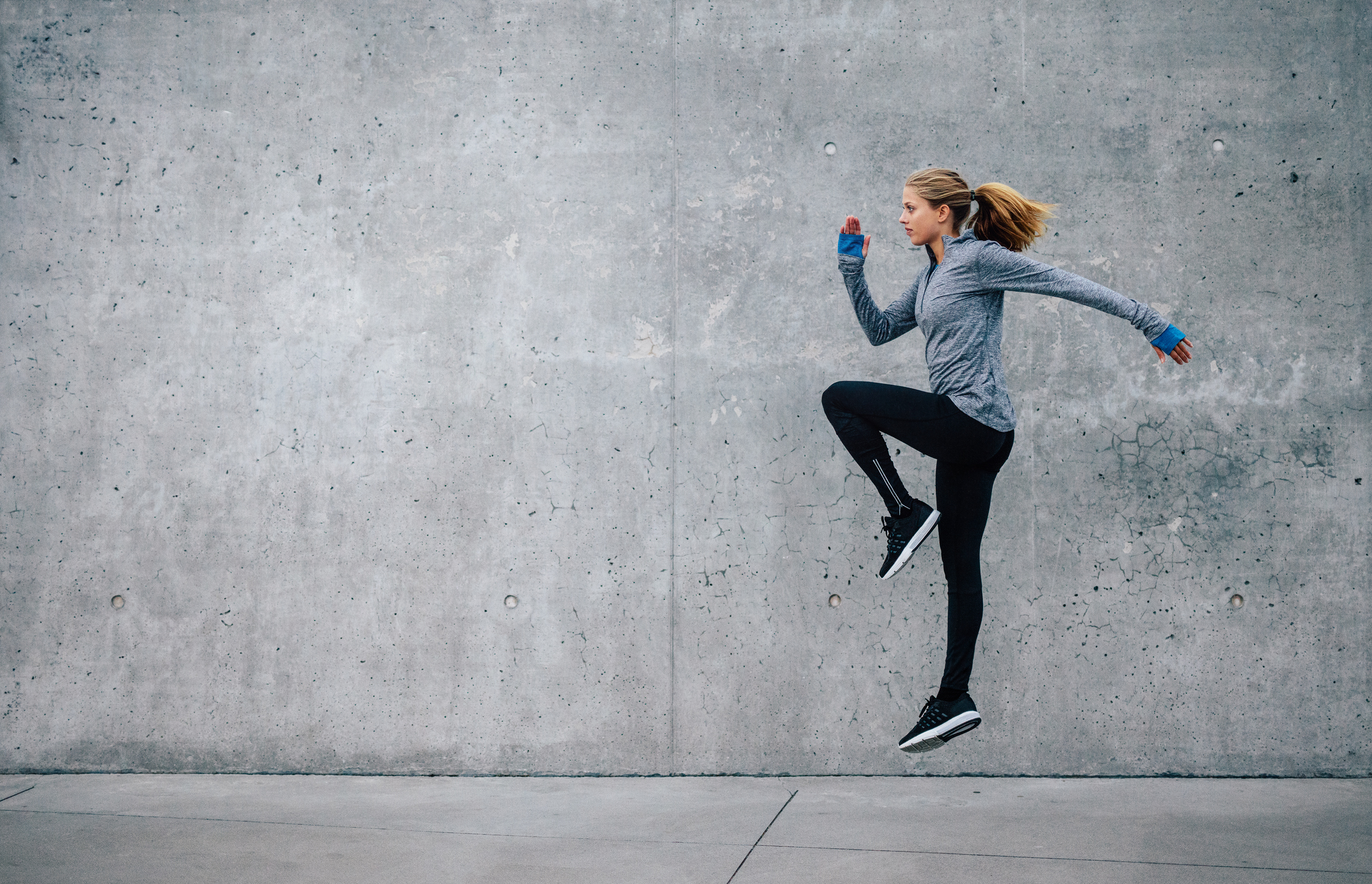 A young woman in athletic apparel works on cardio training beside a cement wall.