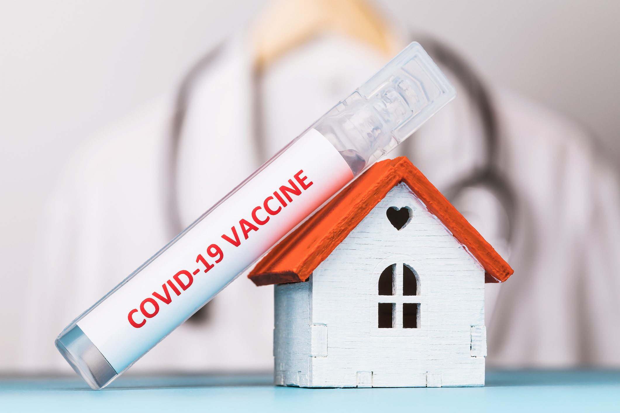 COVID-19 vaccine leaning on top of small toy wooden house with a doctor in the background