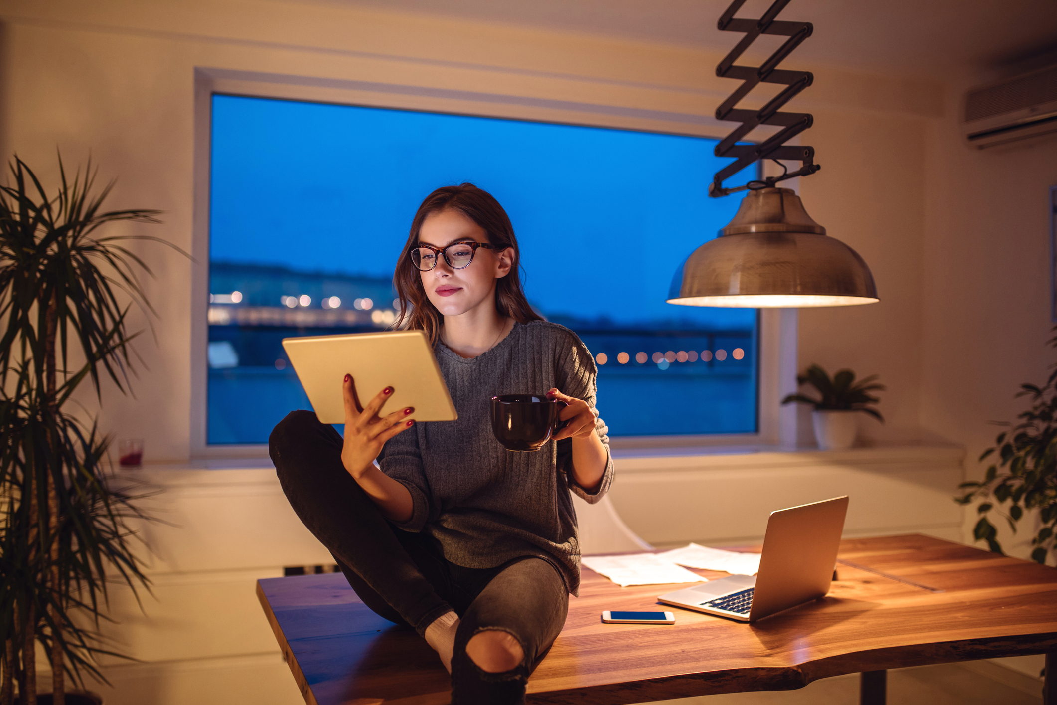 Young woman works from home drinking coffee sitting on a desk and reading from a tablet PC