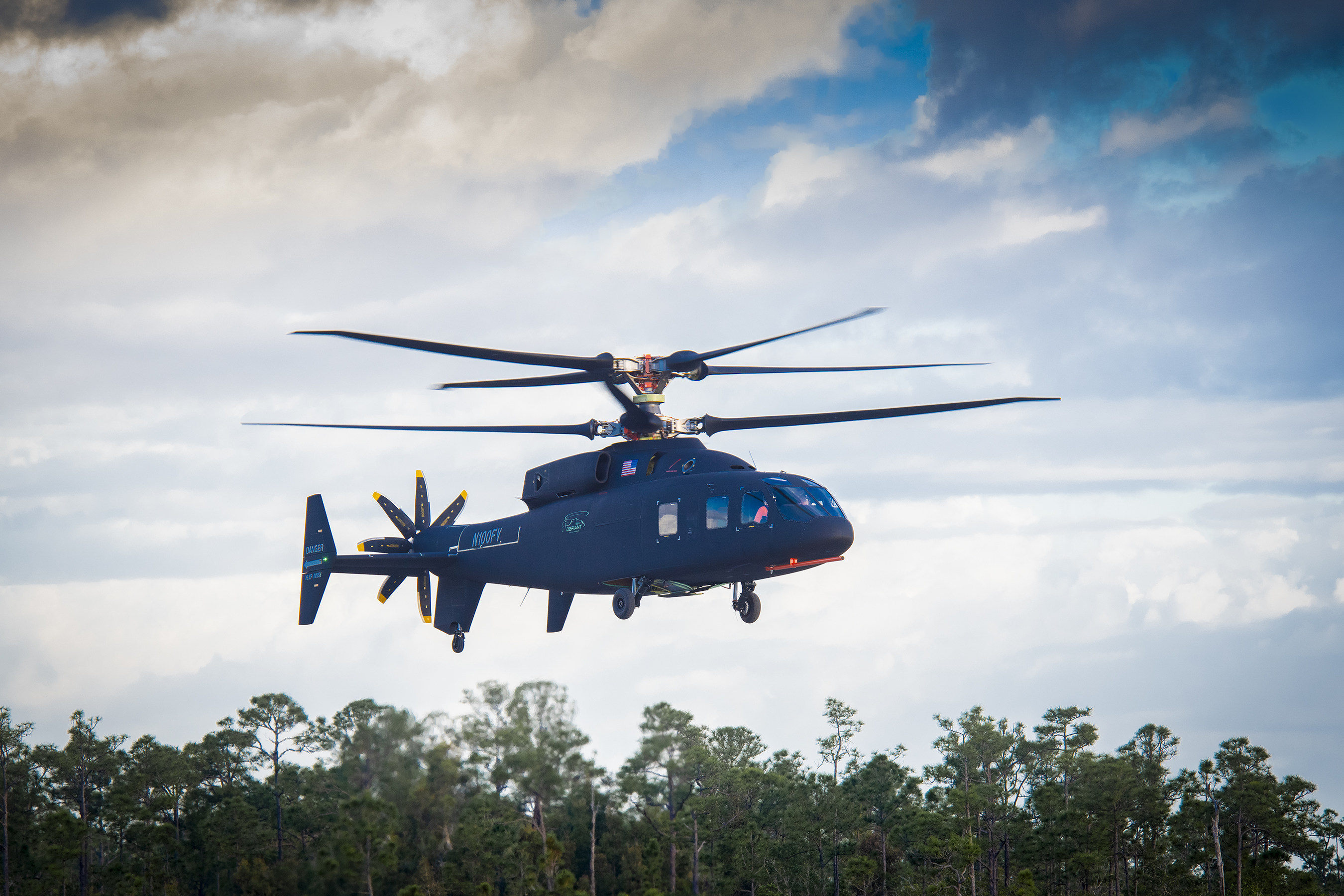 The Defiant hovers over a tree line during its first test flight.