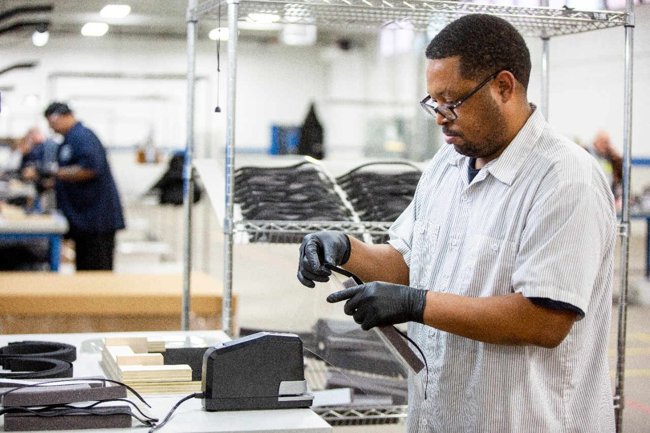 Workers on a well-lit factory floor are shown hand-assembling face shields.