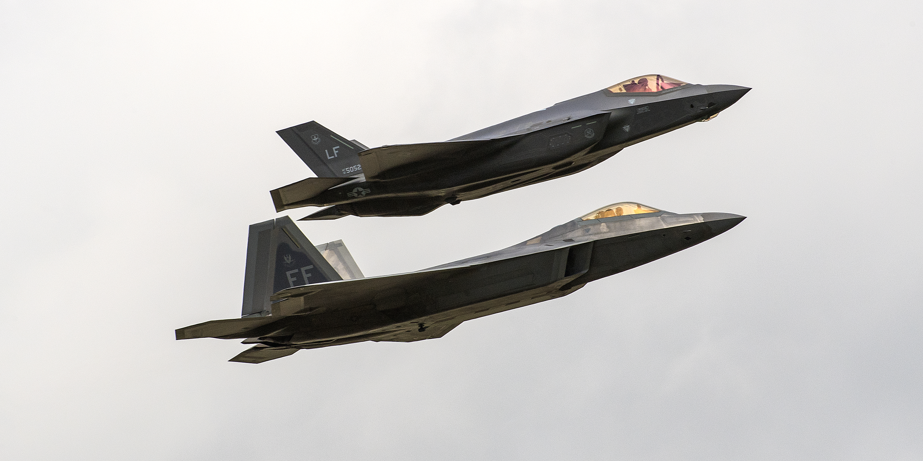 A F-35 and F-22 in position together flying in front of a cloudy sky.