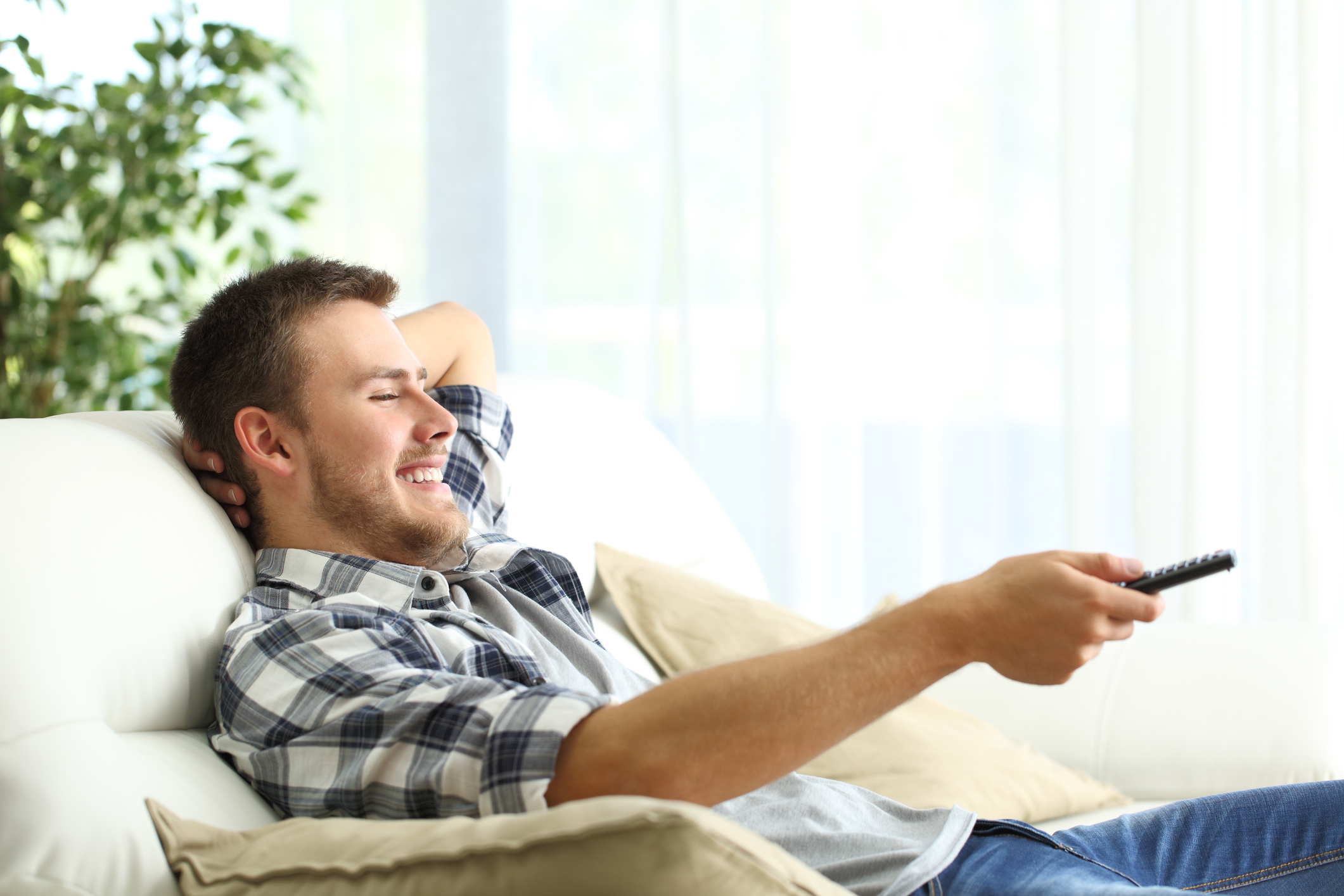 A man on his couch smiling while pointing a remote at his television.