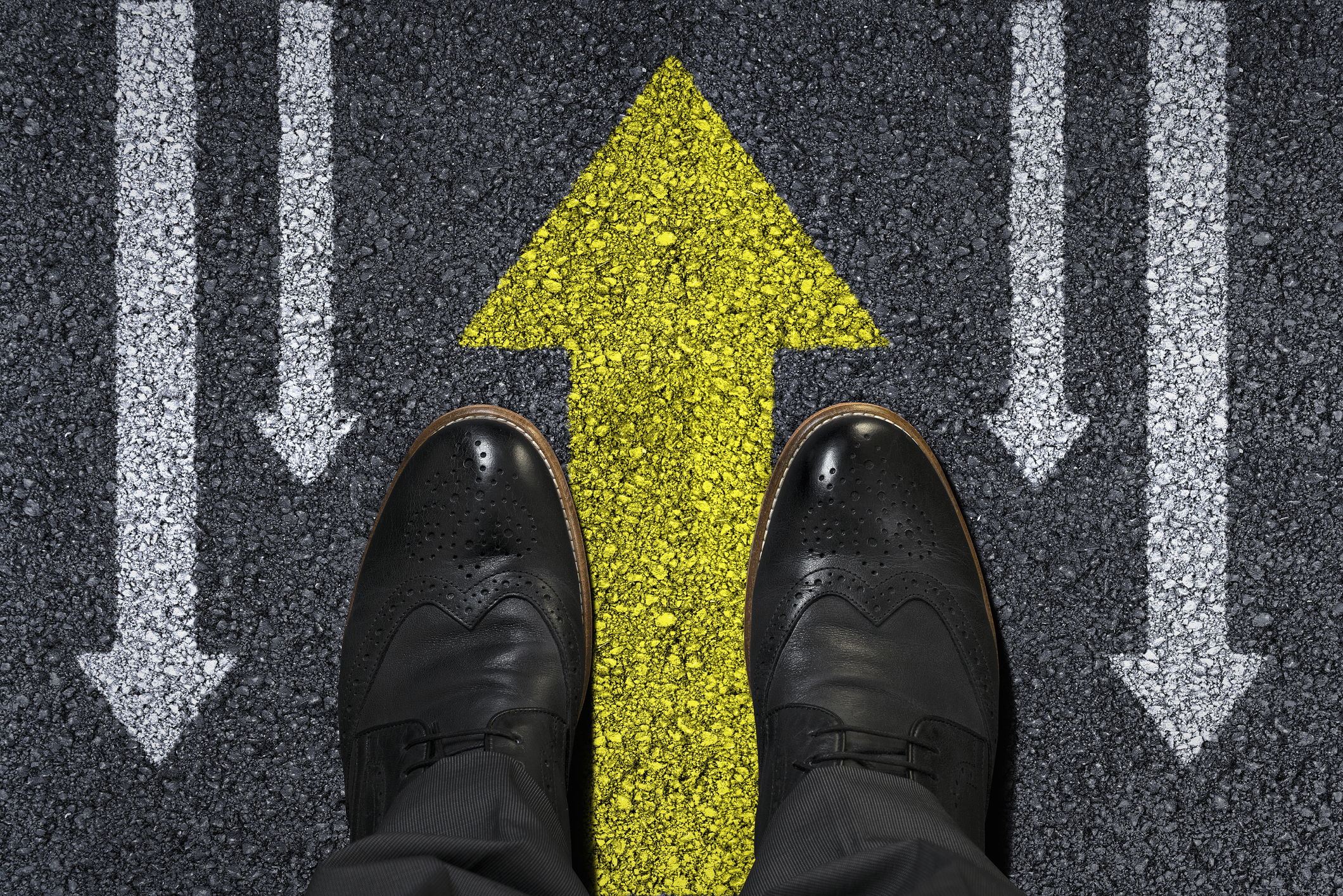 Overhead view of a man's dress shoes straddling a yellow arrow pointing up with white arrows on the sides pointing down
