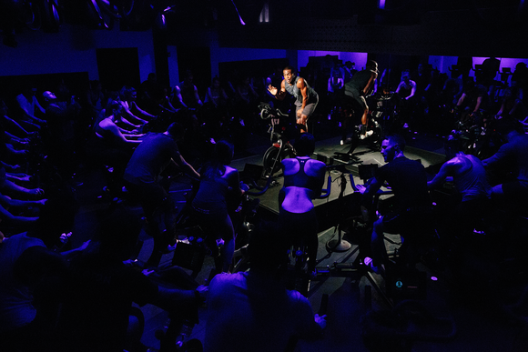 An instructor on a stationary bicycle leads a Peloton group fitness class in purple mood lighting.