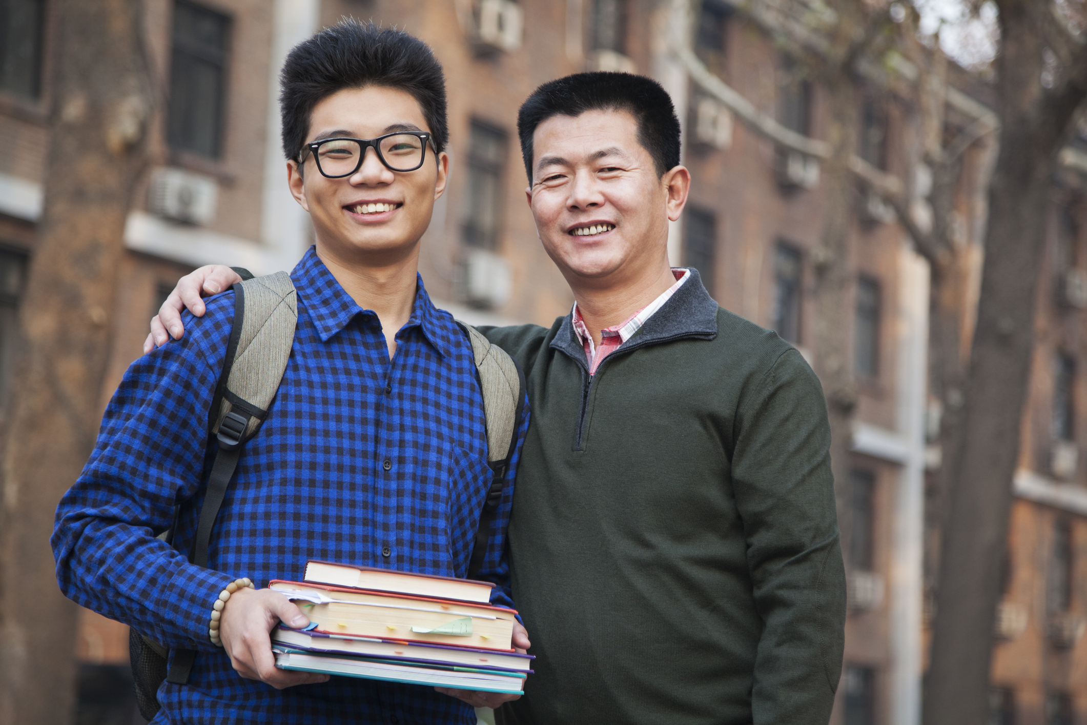 A father with his arm around his son while in front of college dormitories.