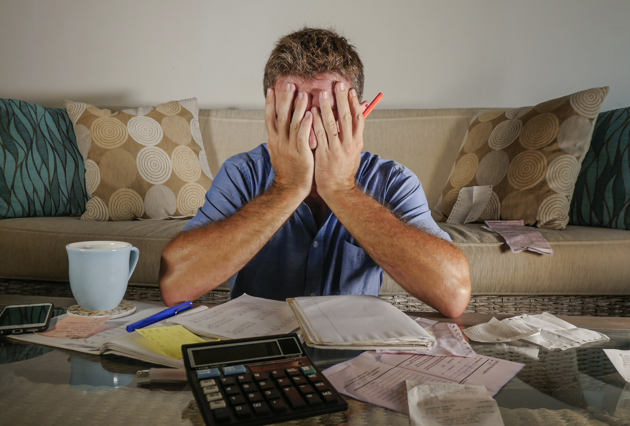 Man covering face, resting elbows on table with calculator and papers spread out