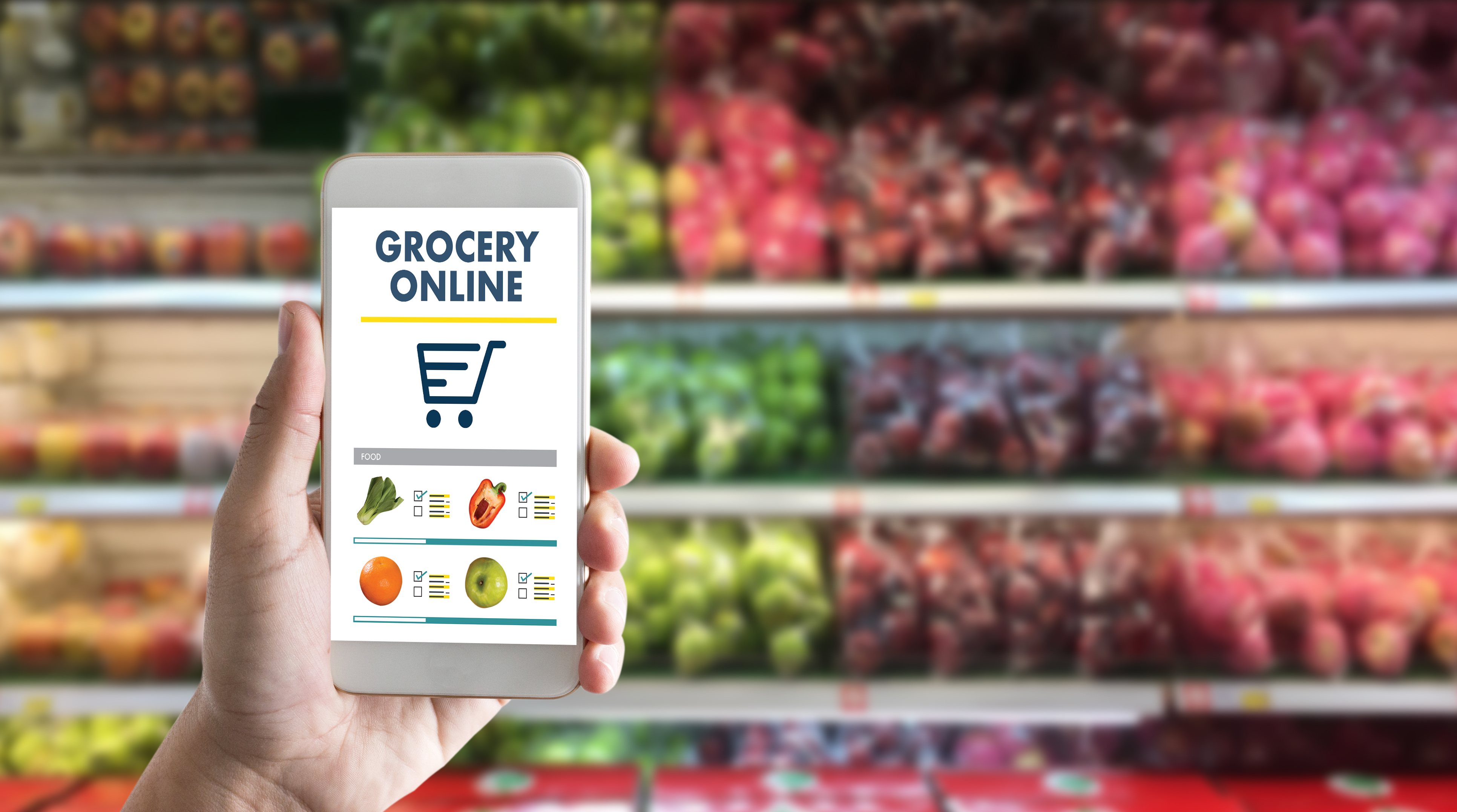 A hand holds a cell phone with "grocery online" written on the screen. Shelves of apples are seen in the background.