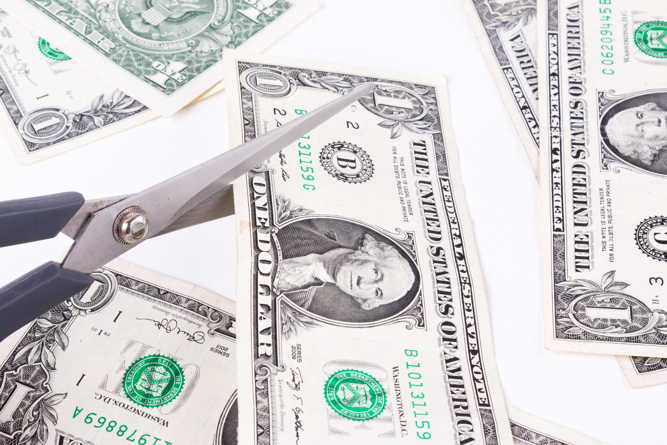 Scissors cutting dollar bill on a table with other bills.