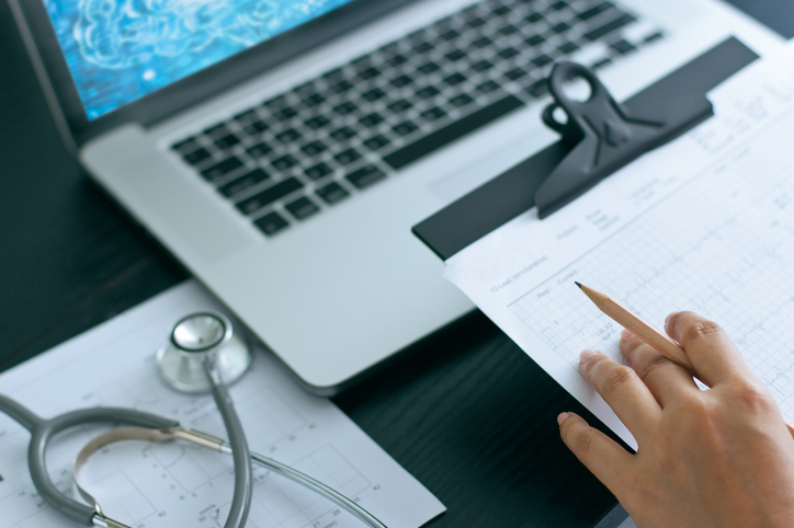 Someone evaluating notes on a clipboard with a laptop and stethoscope present.
