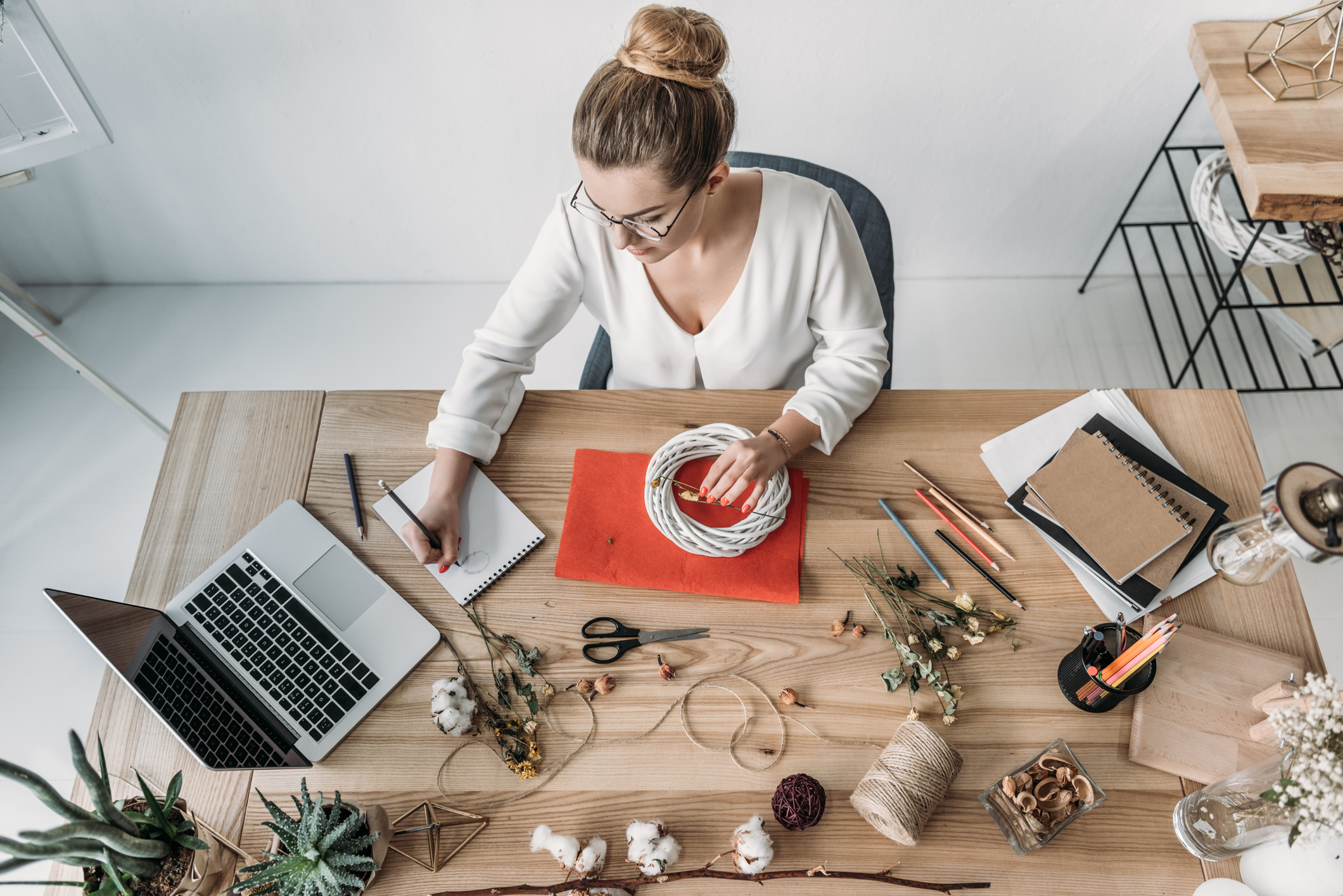 A woman at a worktable with a laptop creates a handmade object.