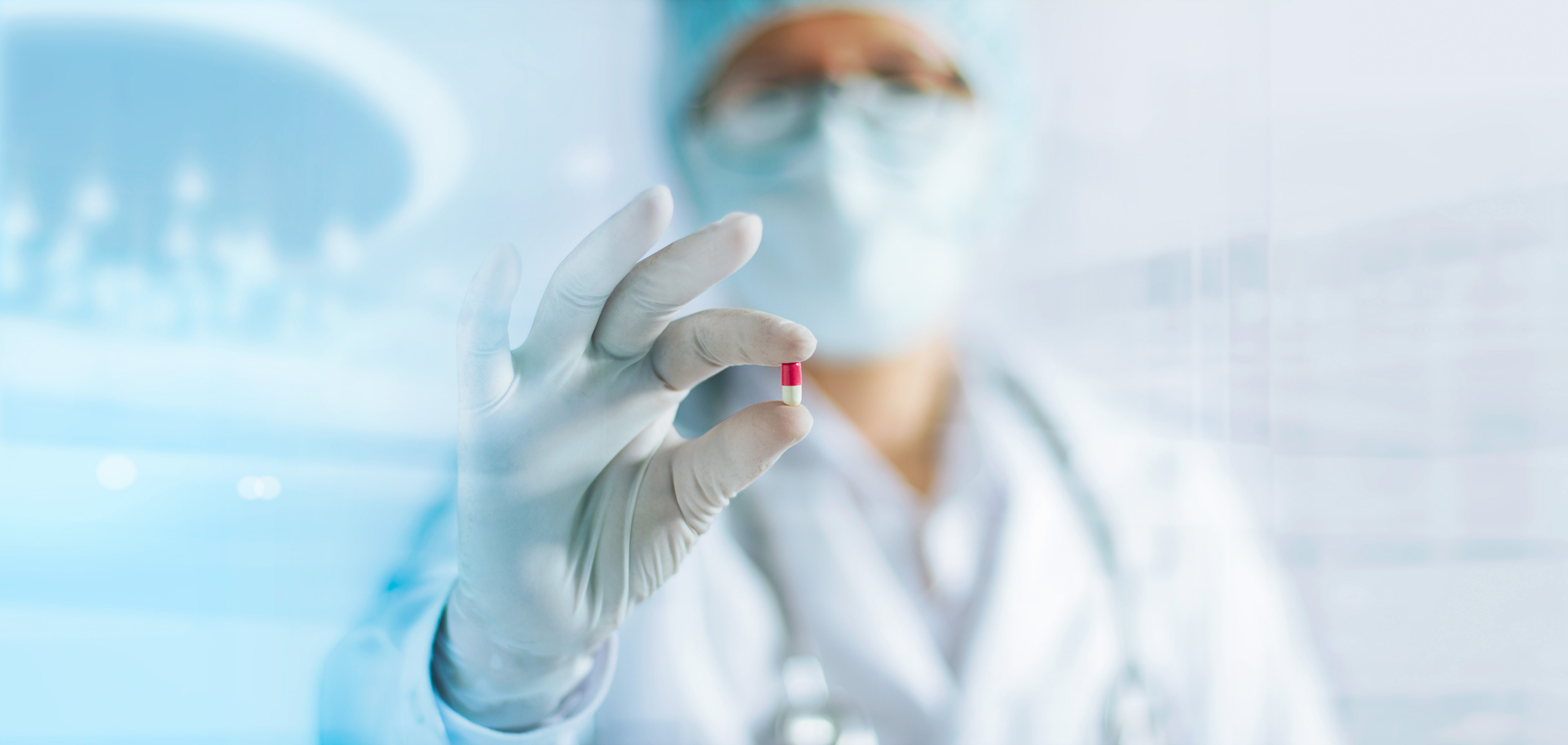 Person in lab coat, safety goggles, and face mask holding a pill in their gloved hand.
