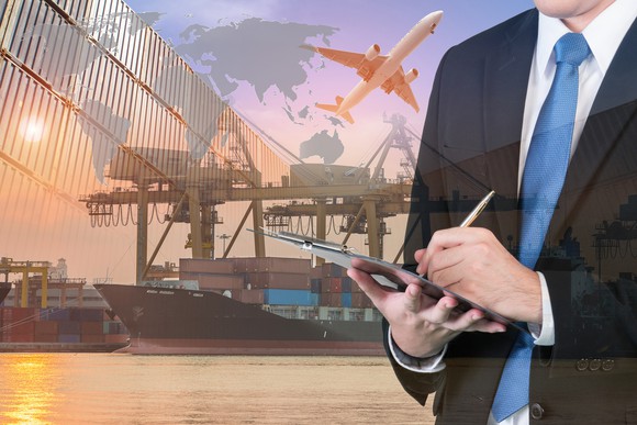 A man in a business suit writing a a clipboard with a container port in the background.