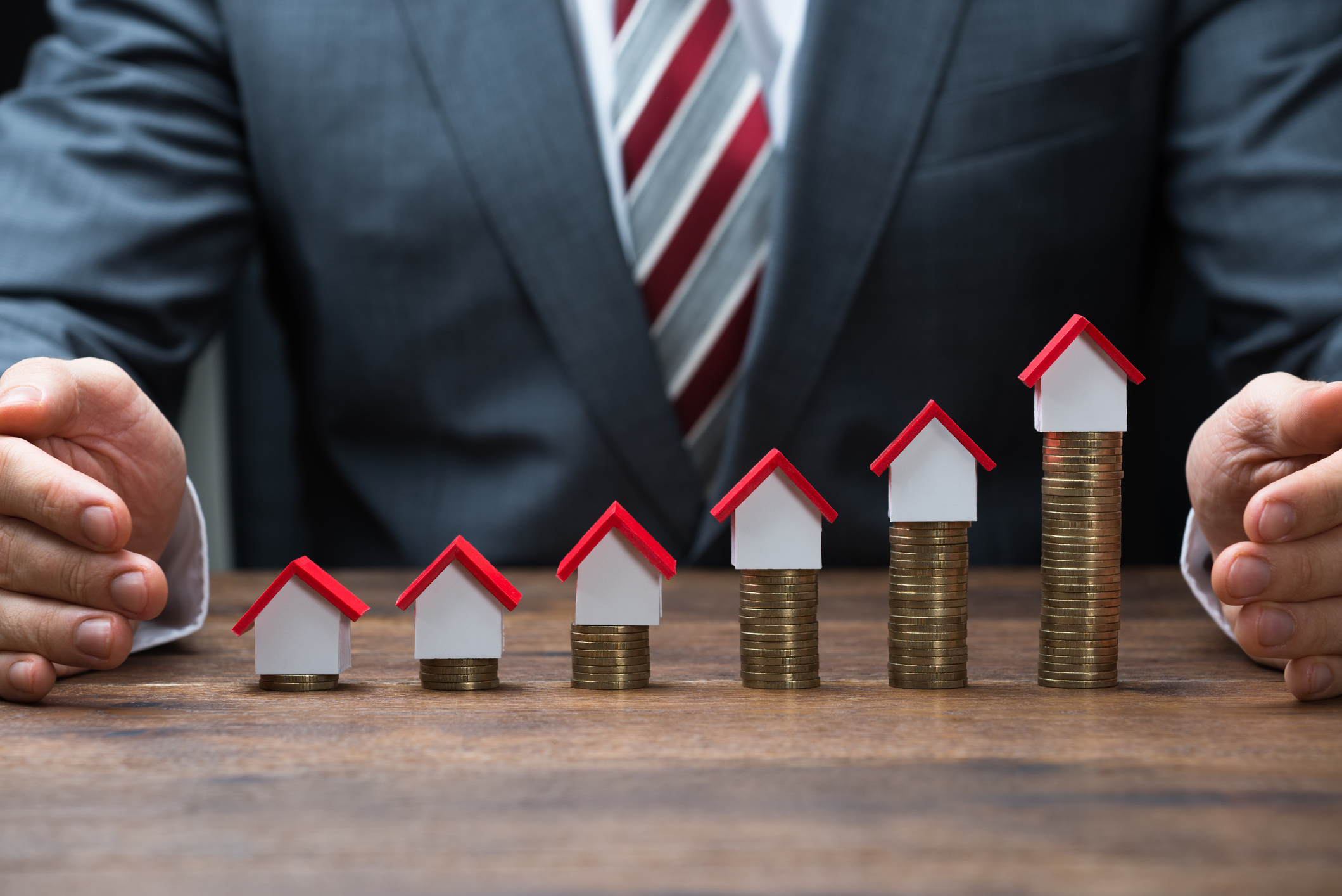 A person in a business suit sitting behind a table that has miniature houses on top of rising stacks of gold coins on it 