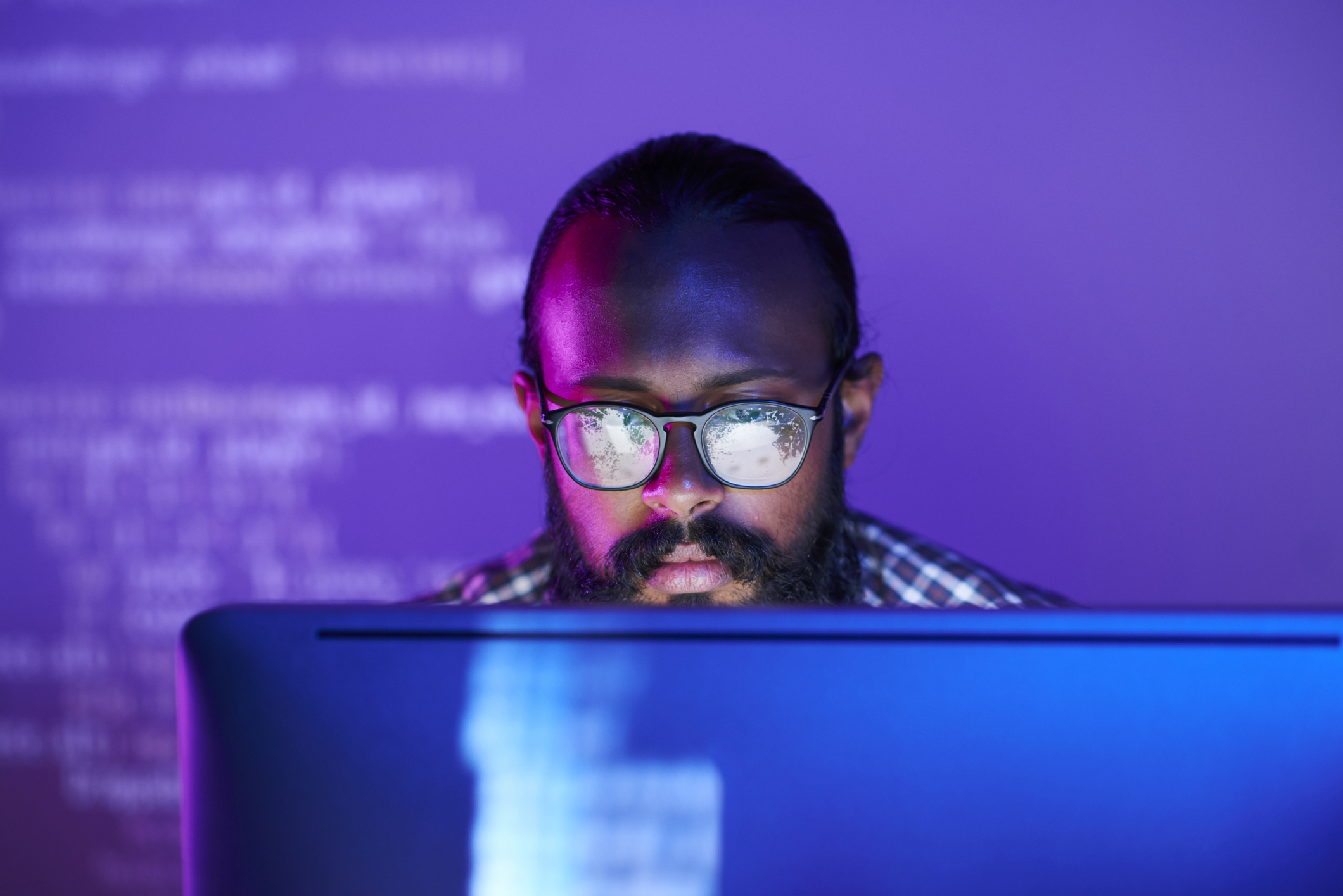 Young man in glasses looks at a desktop screen in a dark room lit with purple light.