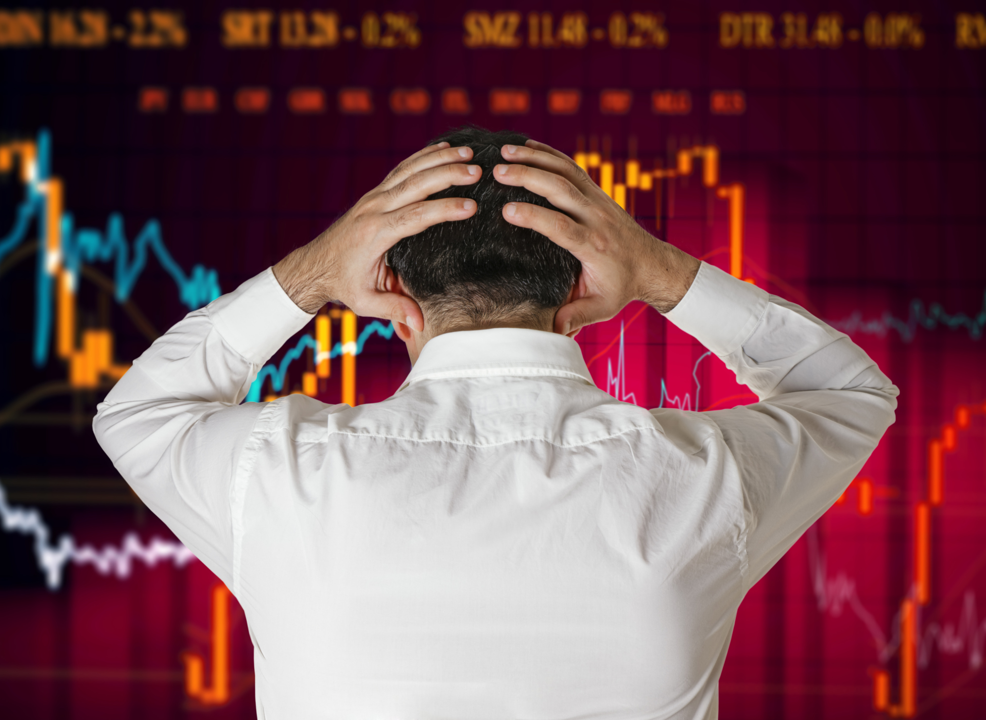 A man places his hands on his head as he stares at a red, downward-pointing stock chart.
