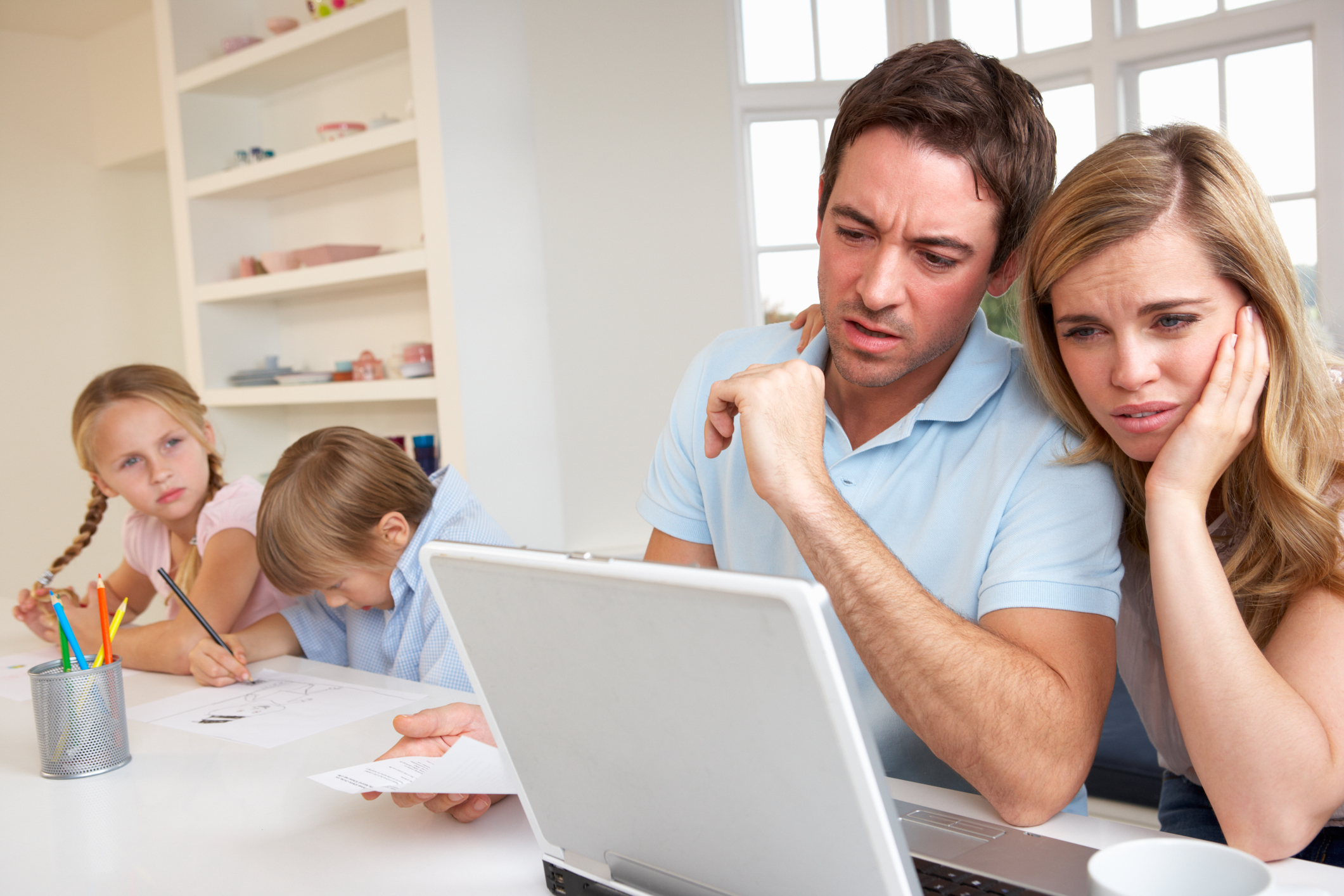 A visibly concerned couple looking at a laptop, with their kids coloring in the background