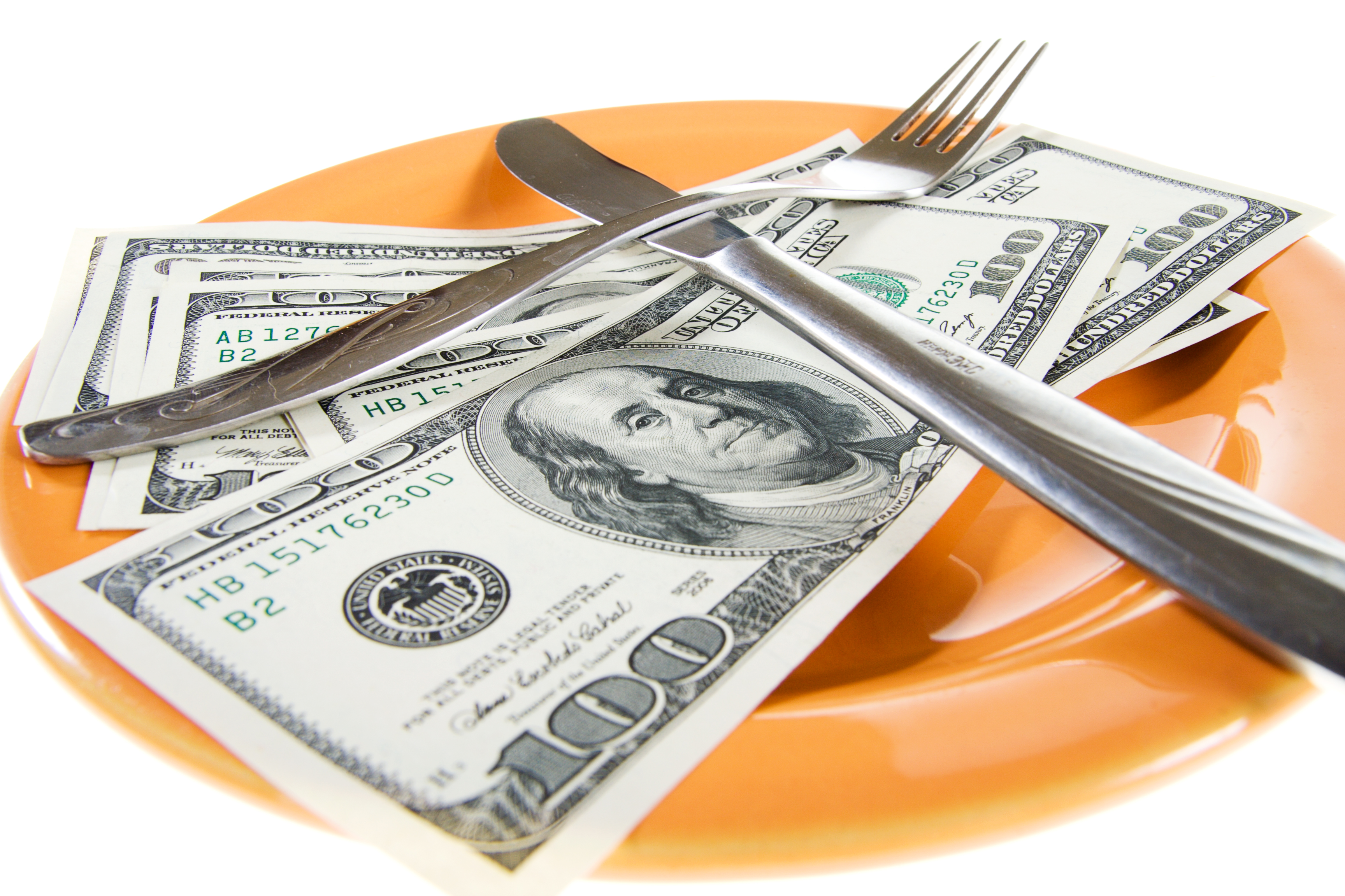 A fork and a knife lie crossed on top of several hundred-dollar bills on an orange plate.