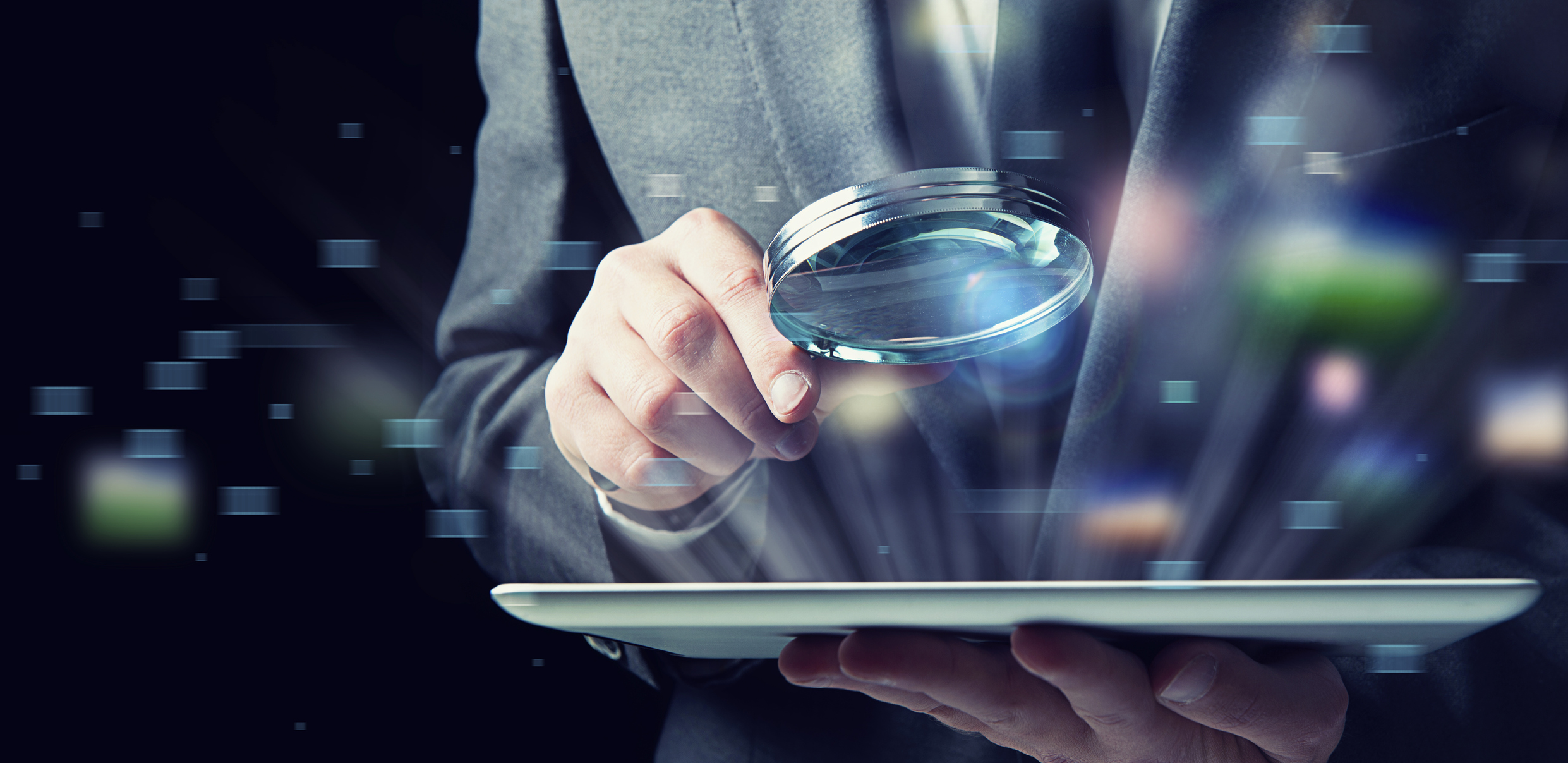 Closeup of a businessman holding a magnifying glass to an electronic tablet.