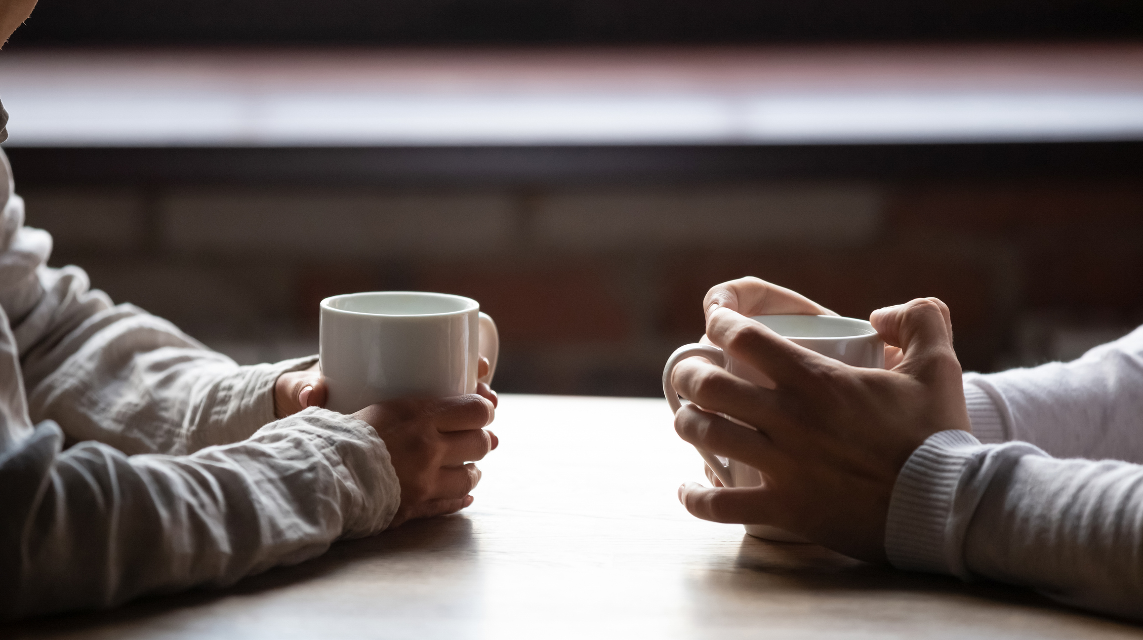 closeup on two pairs of hands holding white cofee mugs on a coffeeshop table.