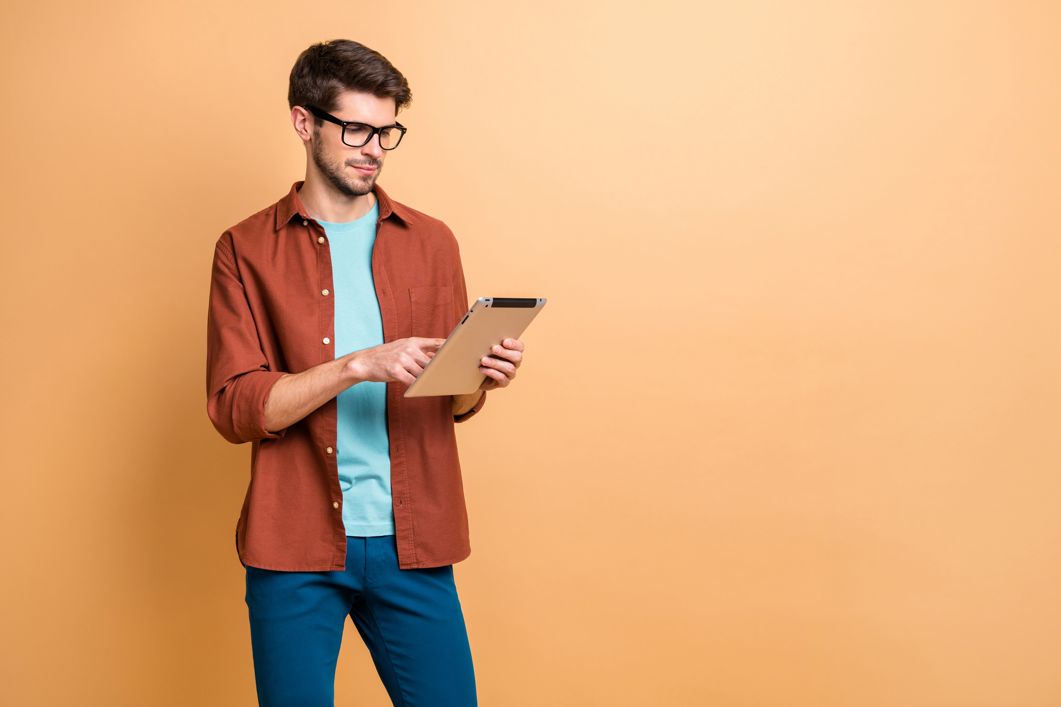 A young man in casual wear and glasses holds and works an electronic tablet.