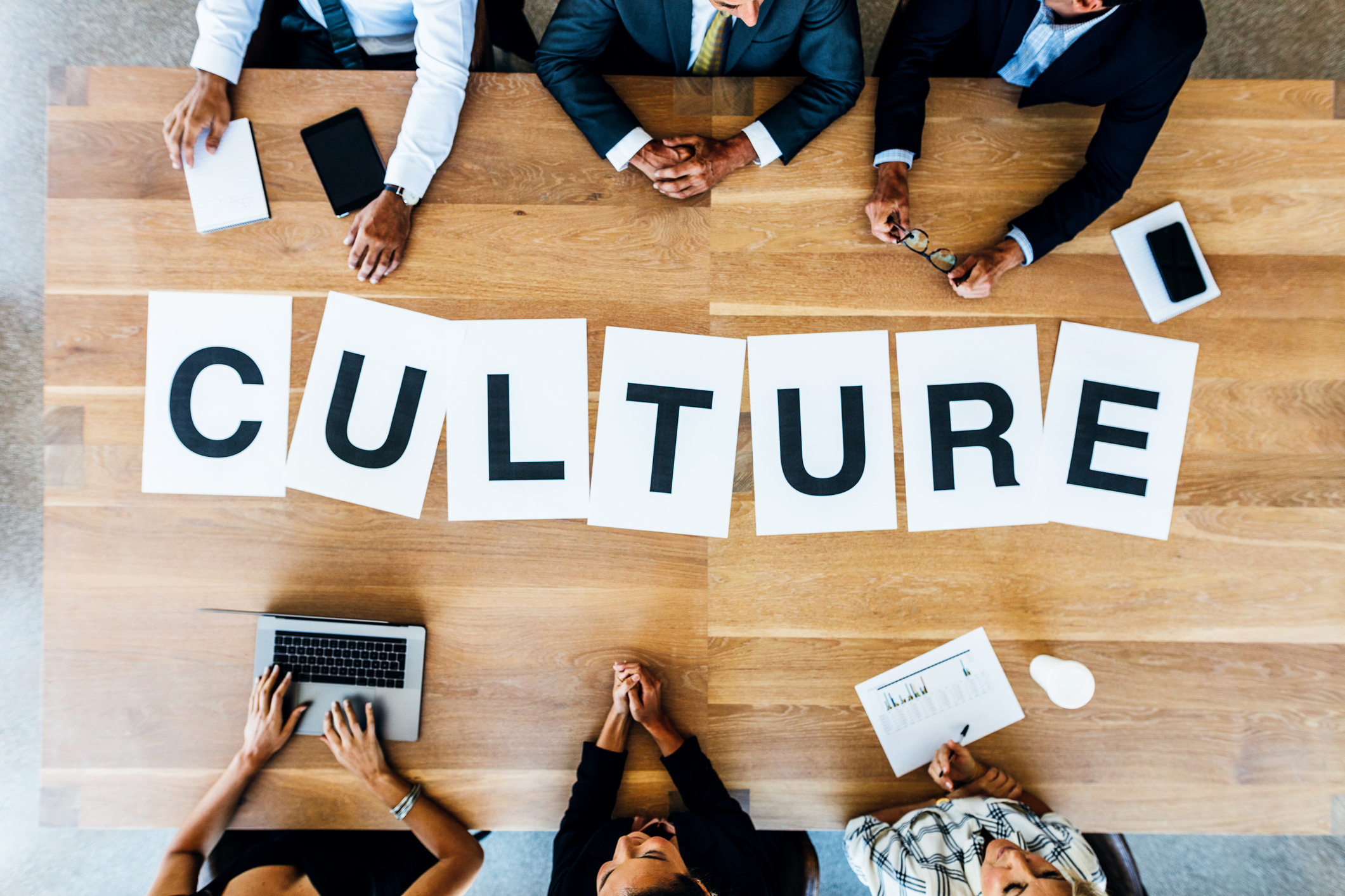 Aerial shot of the word culture printed on one letter per page arranged on a wooden conference table at which six people in business attire sit.