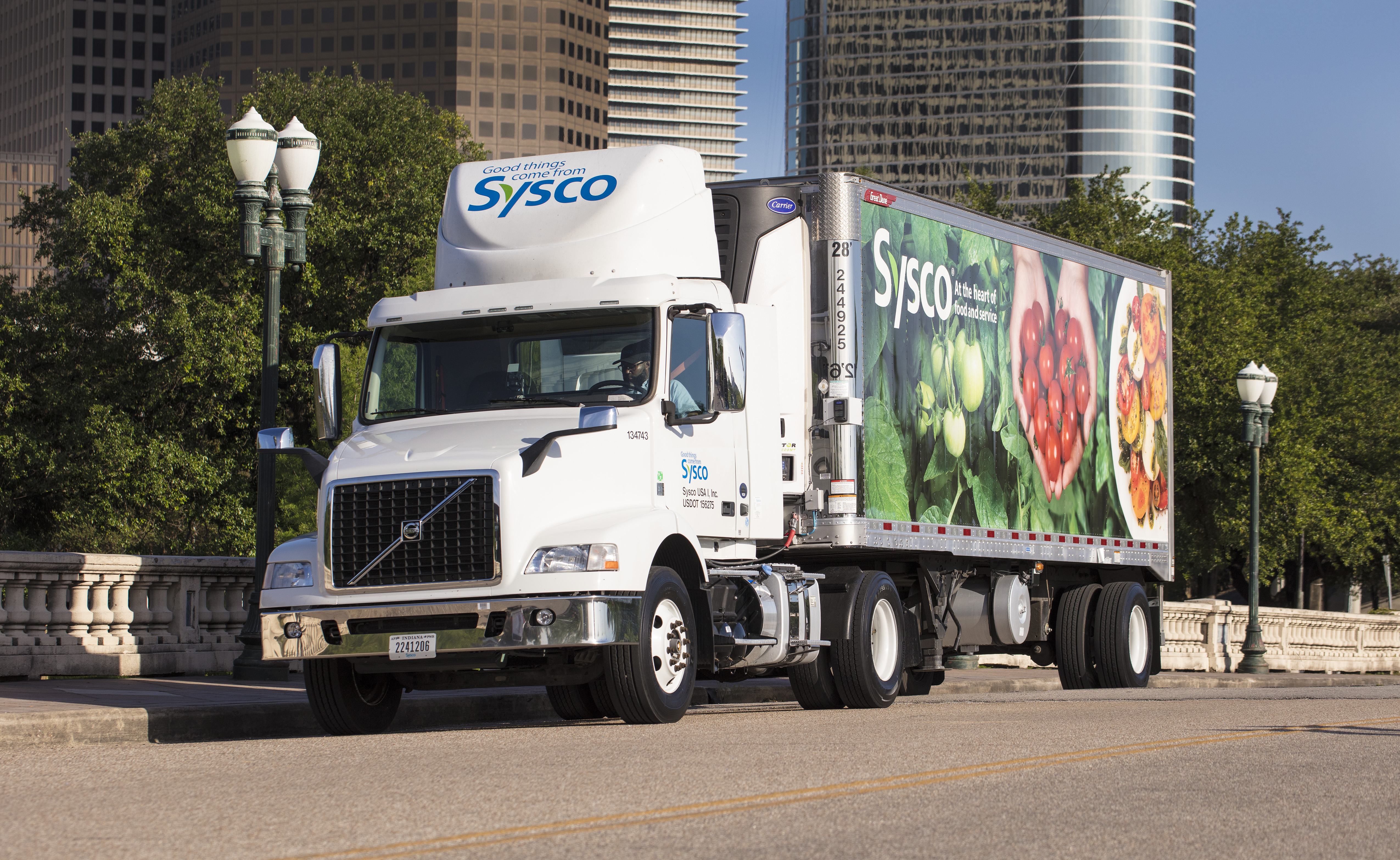 A Sysco delivery truck sits on a roadway in a city with skyscrapers in the background