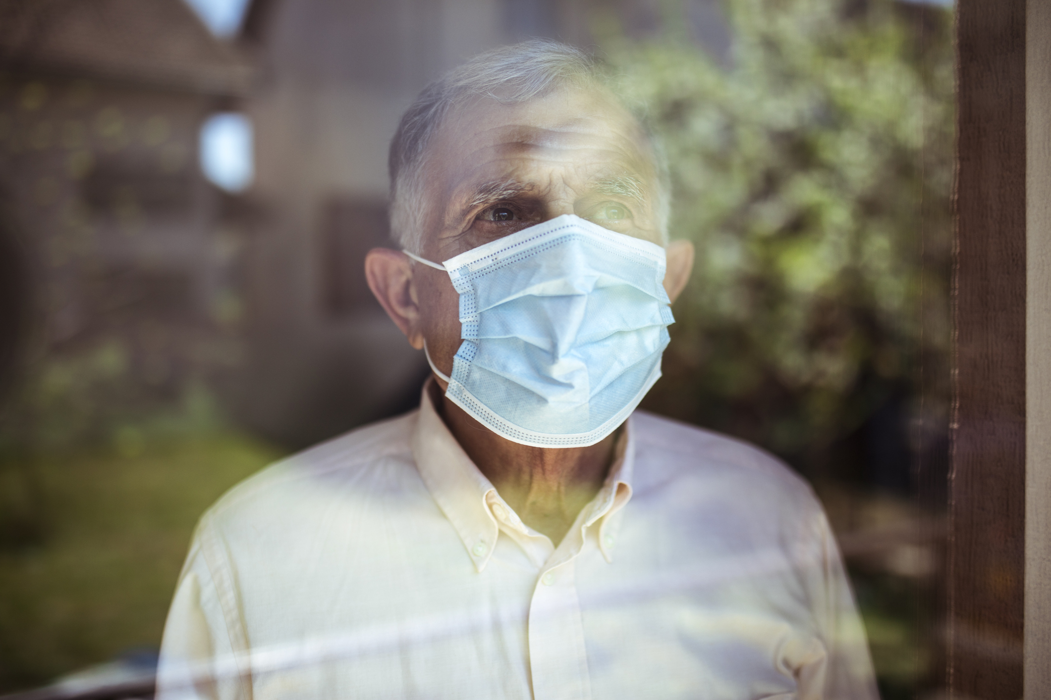 Older man wearing face mask while looking out of a window.